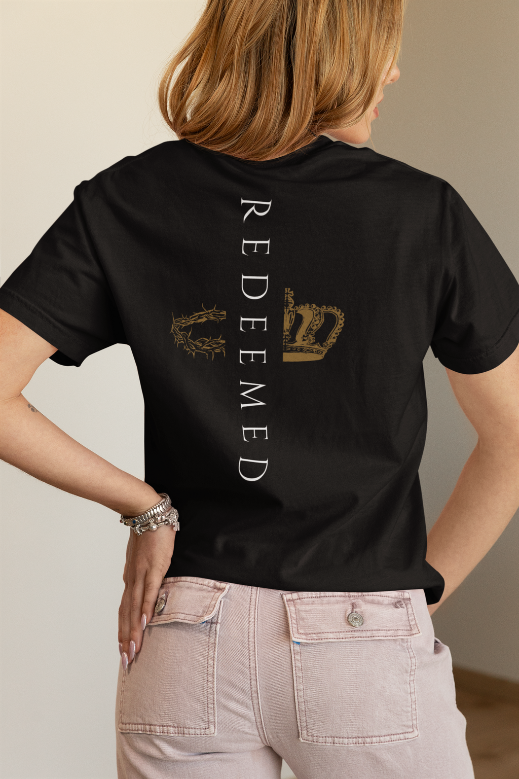 A woman shown from the back wearing a black short-sleeve Christian graphic T-shirt from SeedSowerGraphics with vertical white 'Redeemed' text and a distressed gold crown graphic, paired with light pink jeans.