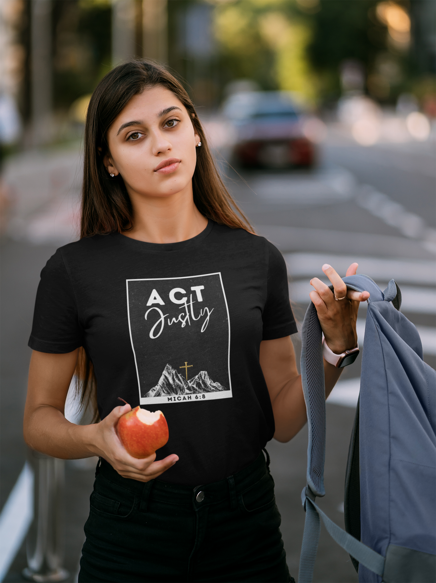 A female student wearing a black Christian graphic t-shirt with the words 'ACT Justly' and a Bible verse printed on the front, holding an apple and a gray backpack in an outdoor street setting.
