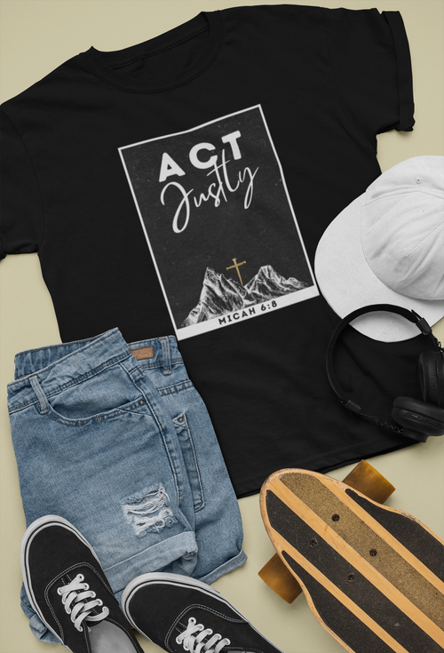 A black Christian graphic T-shirt laid flat with light blue ripped jeans, black sneakers, a white cap, and a skateboard on a beige background. The shirt has a white rectangular graphic with the words “ACT Justly” above a mountain illustration and “Micah 6:8” at the bottom.