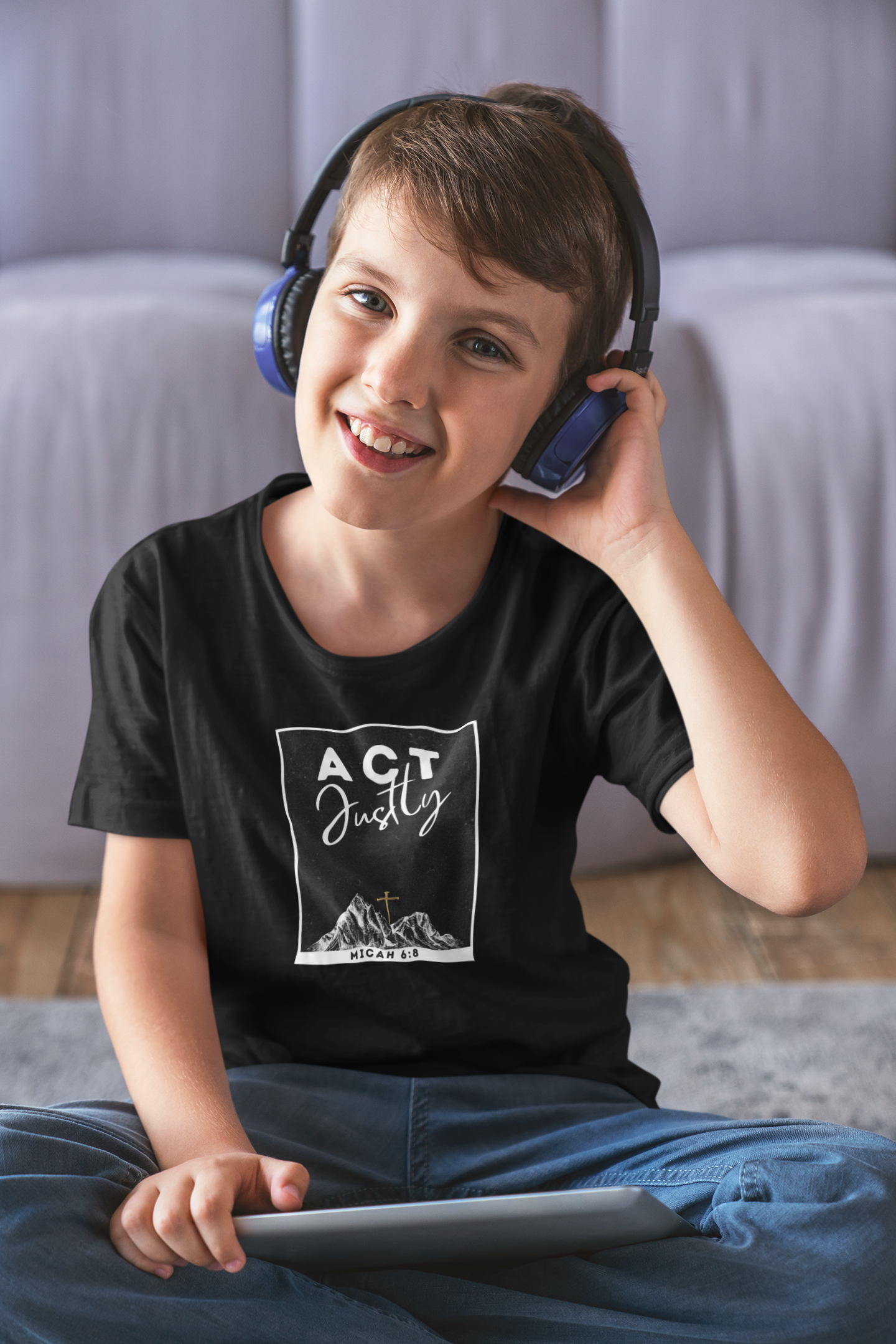 A smiling young boy wearing over-ear headphones and a black Christian graphic t-shirt with the words 'ACT Justly' and a mountain design, sitting on a gray rug in front of a light gray sofa.
