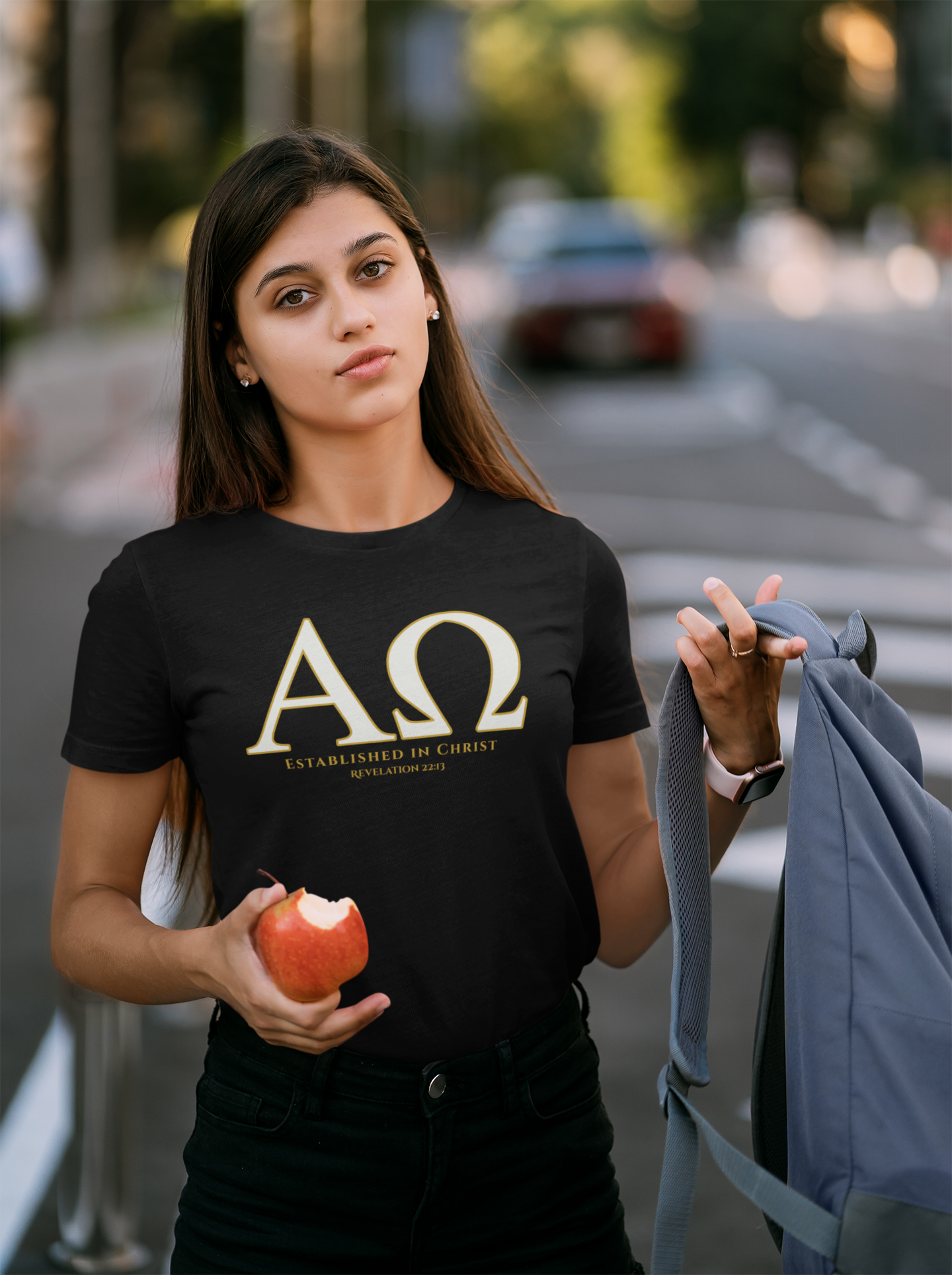 A young woman standing outdoors on a street holding a red apple and a gray backpack, wearing a black faith based t-shirt with a large gold Greek letter 'ΑΩ' and the text 'ESTABLISHED IN CHRIST REVELATION 22:13' printed on the front.