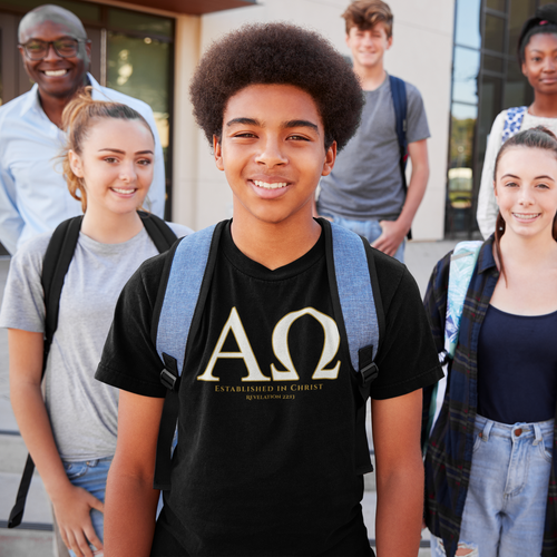 A smiling young man in the center wears a black Christian graphic T-shirt with large Greek letters Alpha and Omega, with the words "Established in Christ' Revelation 22:13', and blue-gray backpack straps, standing outdoors with four classmates in front of a modern building.