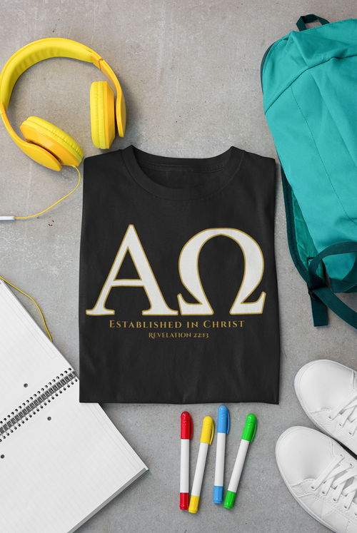 A black Christian graphic T-shirt laid flat on a gray surface, surrounded by yellow headphones, a teal backpack, a notebook, four colored markers, and white sneakers. The shirt has large gold Greek letters 'Αlpha/Omega' with smaller gold text reading 'ESTABLISHED IN CHRIST' and 'REVELATION 22:13'.