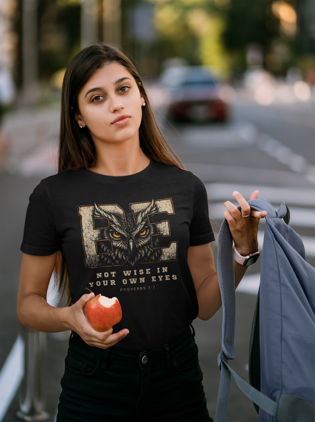 A young woman standing outdoors on a blurred street background, wearing a black Christian graphic T-shirt with a large owl design and the text 'Be NOT WISE IN YOUR OWN EYES PROVERBS 3:7'. She is holding an apple in one hand and a gray backpack in the other.