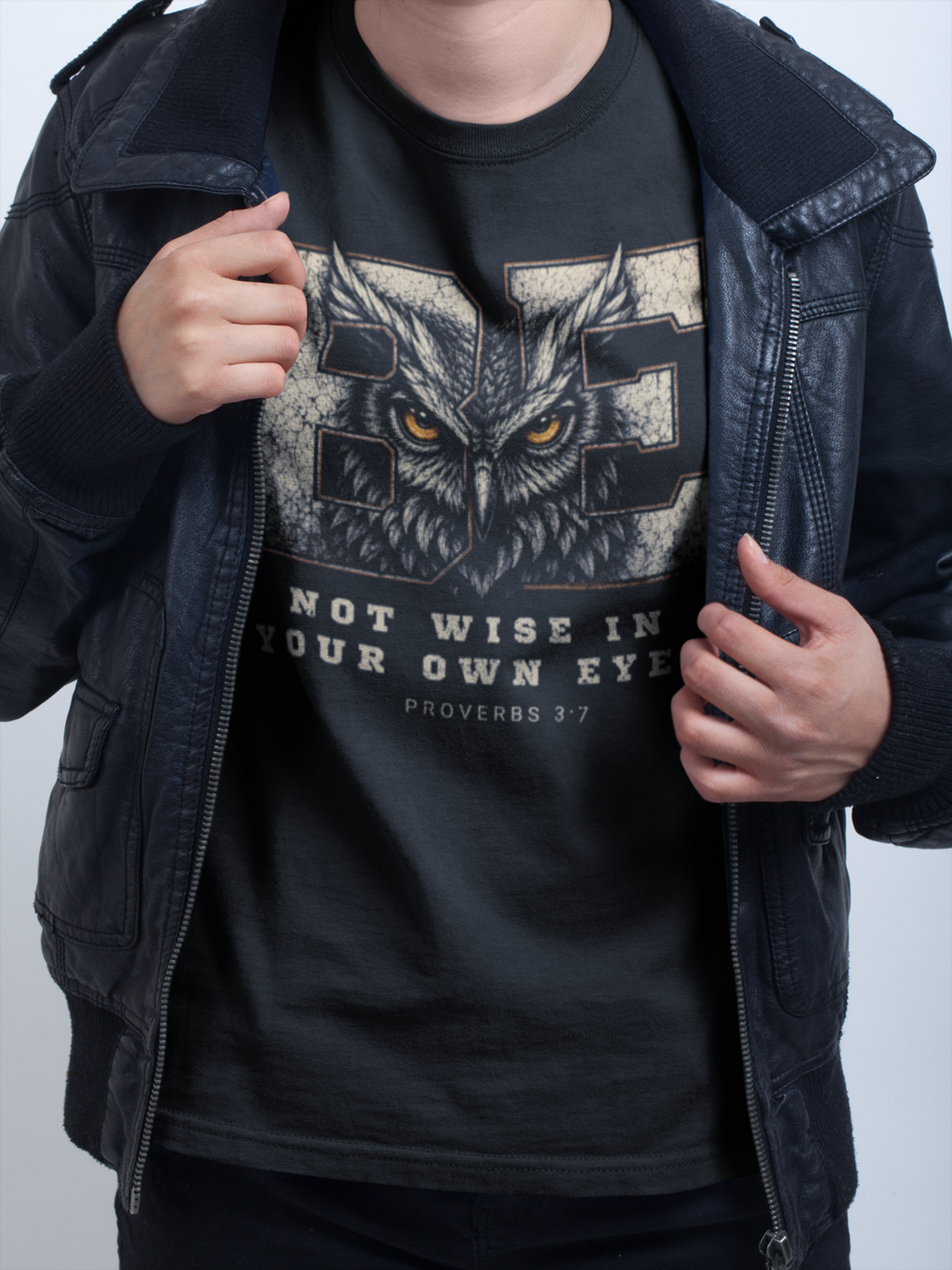 A person wearing a black Christian apparel t-shirt with an owl design and the text 'BE NOT WISE IN YOUR OWN EYE PROVERBS 3:7', layered under an open black leather jacket. 