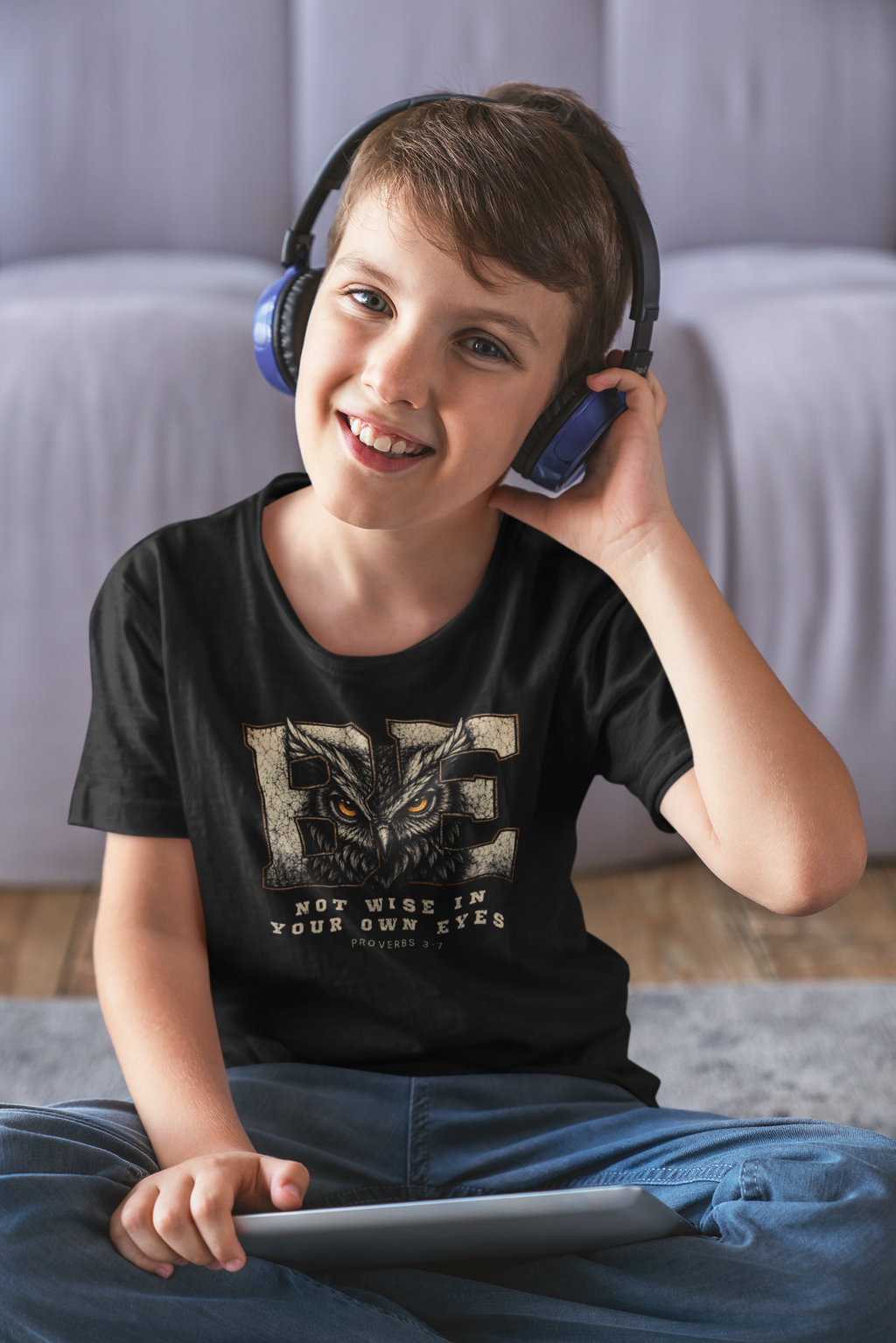 A smiling young boy wearing over-ear headphones and a black Chrisitan graphic t-shirt with an owl design, sitting cross-legged on a gray rug in a living room setting.