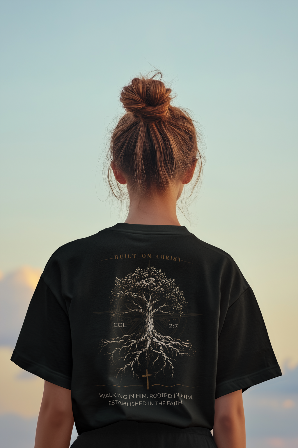 A girl viewed from behind wearing a black Bible verse t-shirt with a detailed white graphic of a tree with roots and text on the back, set against a soft sky background.