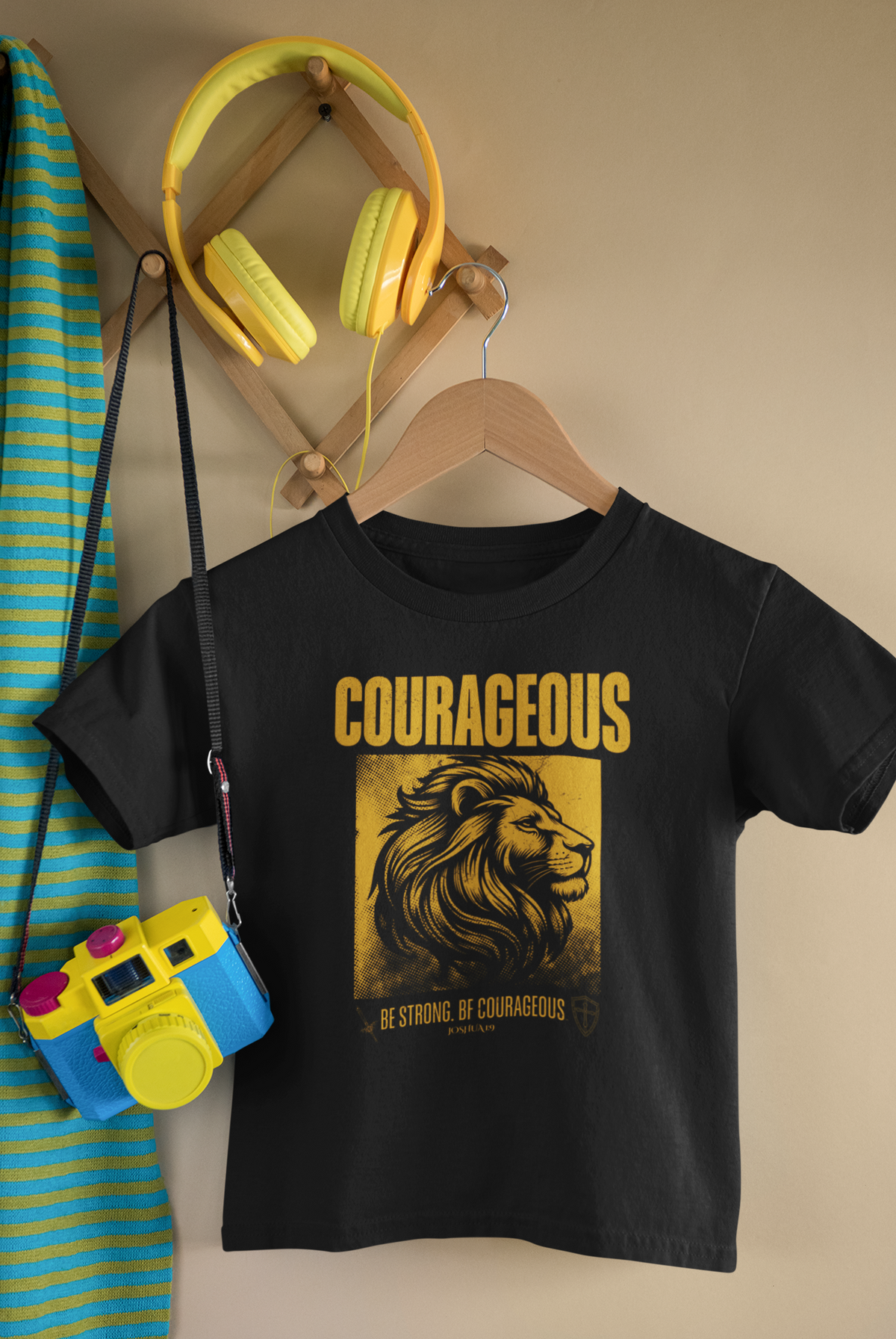 A black short-sleeve Christian graphic T-shirt hanging on a wooden hanger against a beige wall. The shirt features a large yellow and gold graphic of a lion's head with the text 'COURAGEOUS' and 'BE STRONG, BE COURAGEOUS' below it.