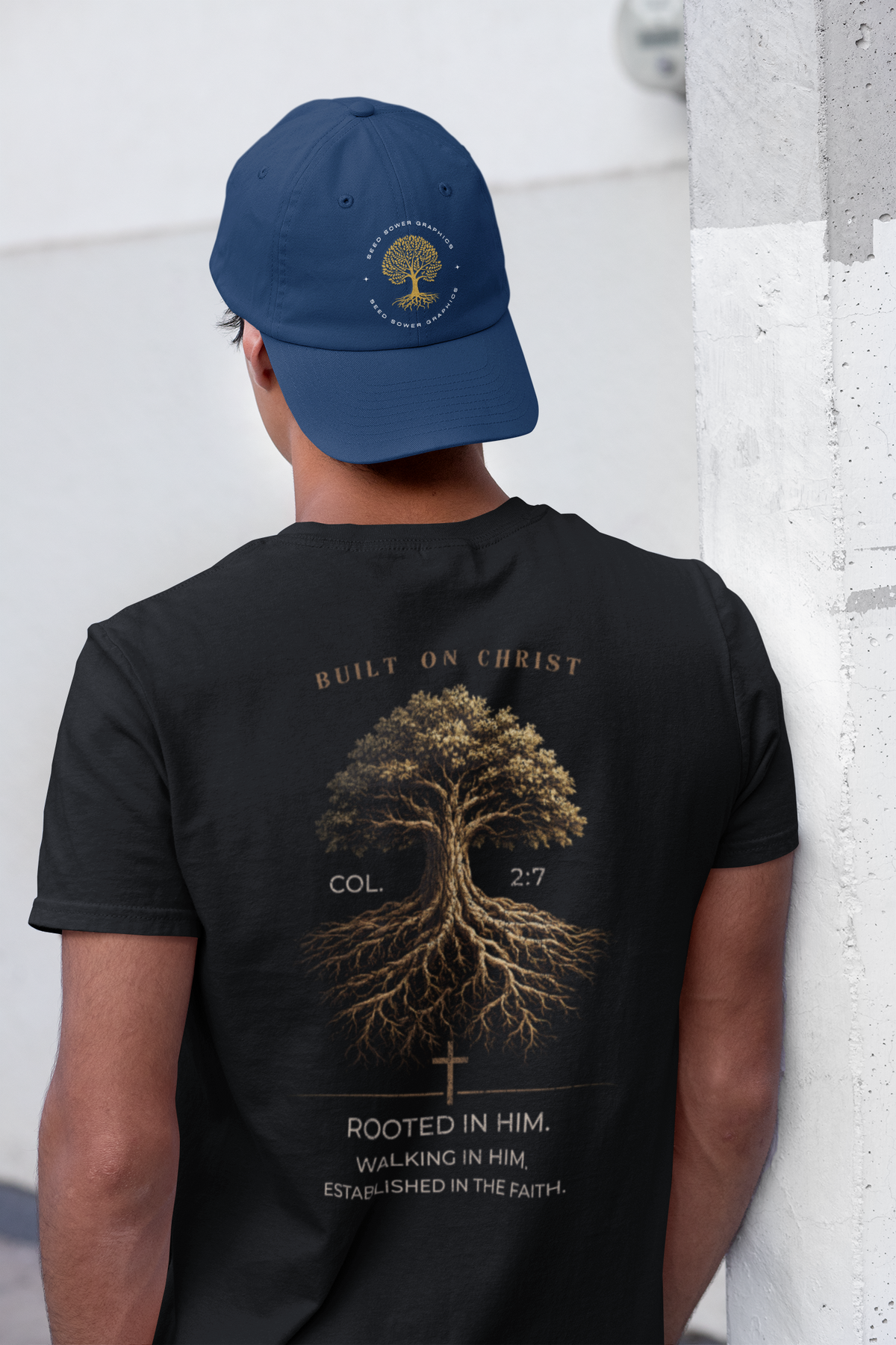 A man viewed from behind wearing a navy blue baseball cap and a black Christian graphic  t-shirt. The back of the t-shirt features a large gold graphic of a tree with roots, accompanied by the text 'BUILT ON CHRIST' and 'ROOTED IN HIM.
