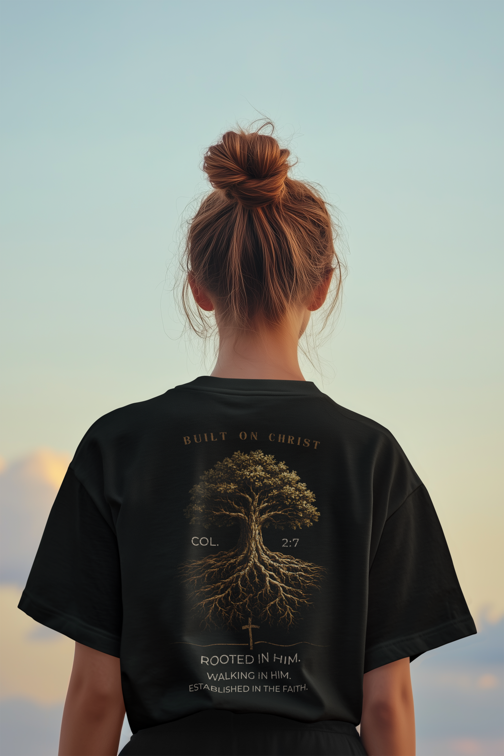 A woman viewed from behind wearing a black Christian graphic T-shirt with a large tree design and text on the back, set against a soft sky background.