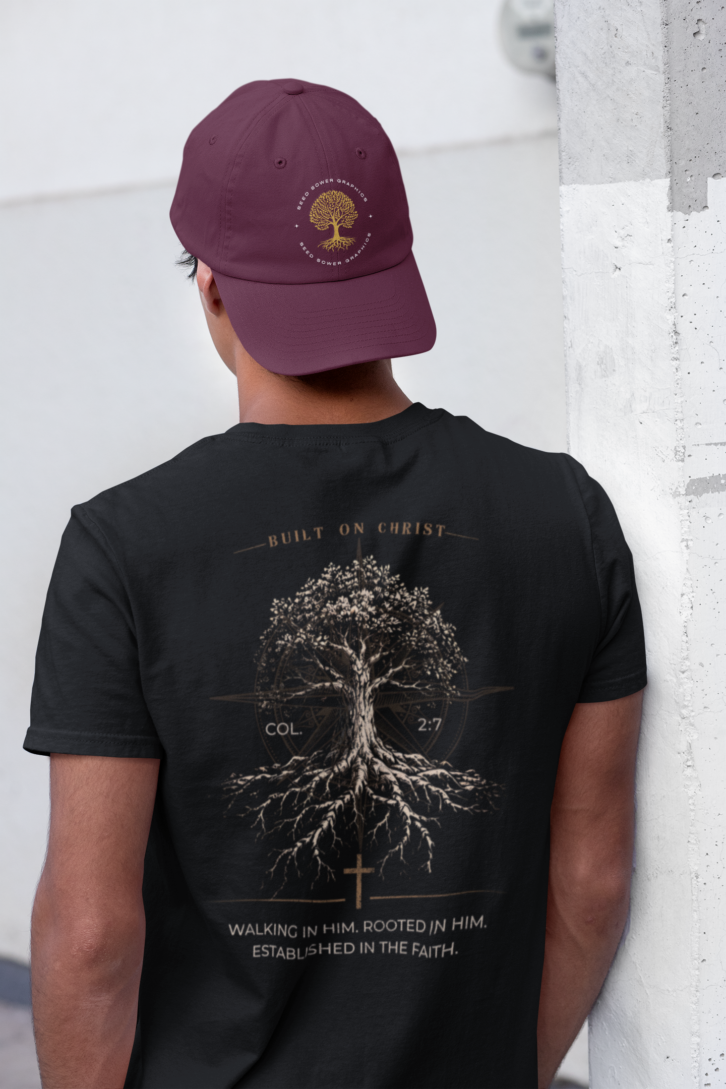 A young man shown from behind wearing a black Bible verse t-shirt with a large graphic of a tree with roots and the text 'BUILT ON CHRIST' and 'WALKING IN HIM, ROOTED IN HIM, ESTABLISHED IN THE FAITH' with Col 2:7. He is also wearing a maroon baseball cap with a gold tree logo that says SeedSowerGraphics.