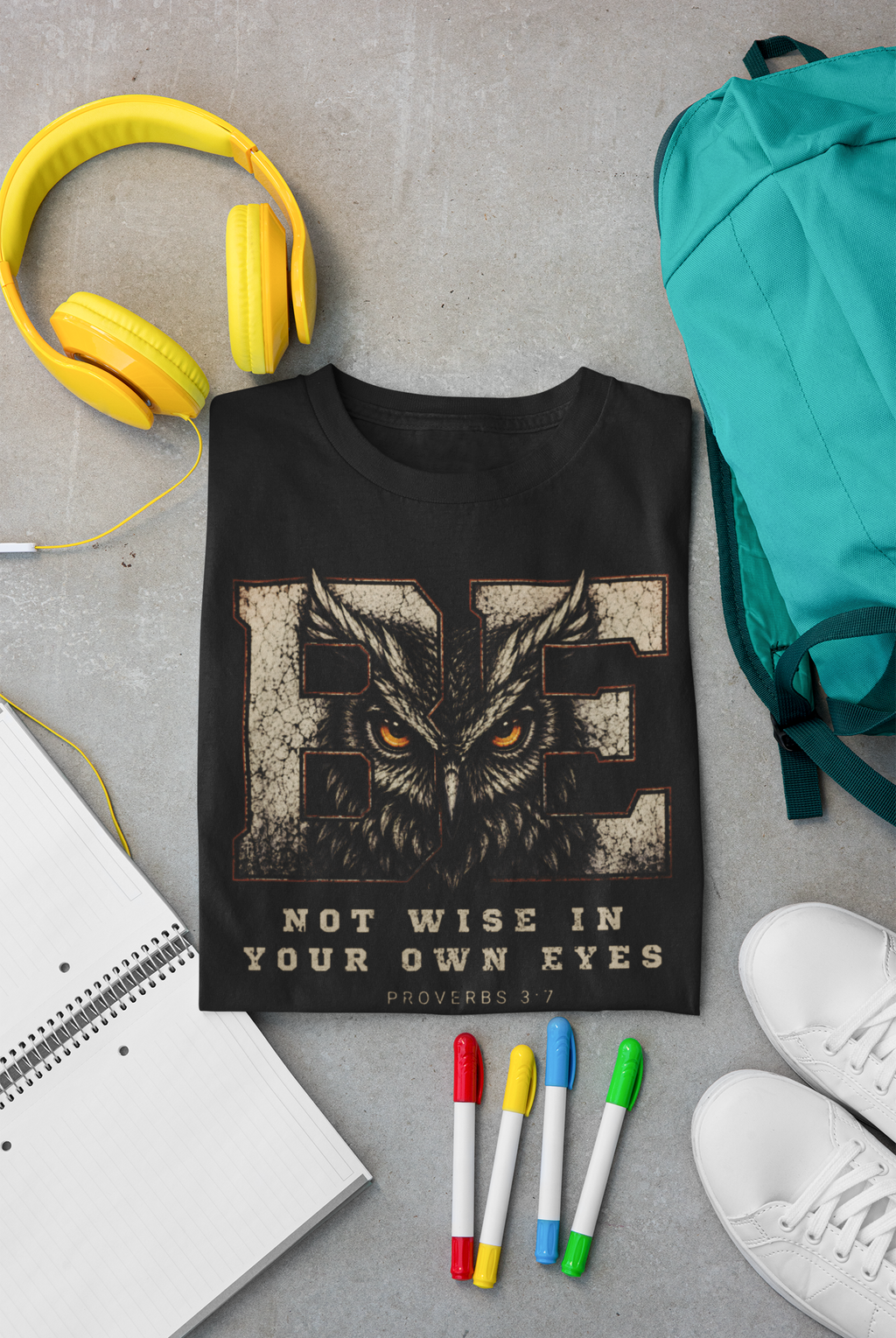 A folded black Christian graphic T-shirt is arranged in a styled flat lay on a gray surface, surrounded by yellow headphones, a teal backpack, white sneakers, colored markers, and a notebook. The shirt features a large owl graphic over distressed beige letters with the text 'BE NOT WISE IN YOUR OWN EYES' and 'PROVERBS 3:7'.