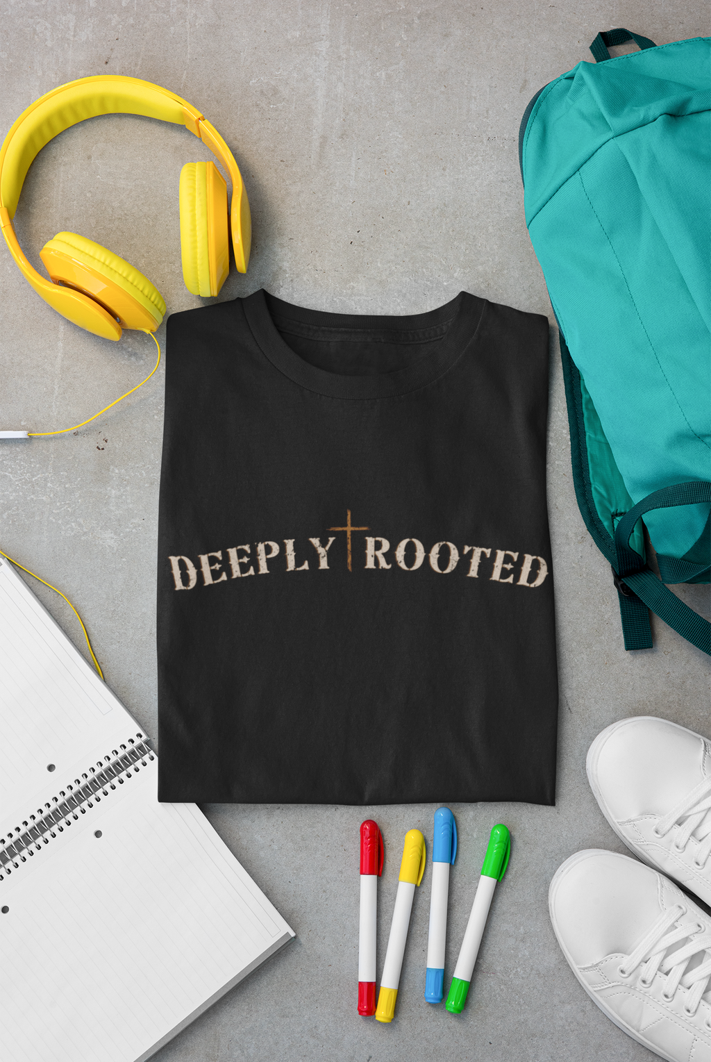 A folded black Christian graphic T-shirt with the words 'DEEPLY ROOTED' and a cross symbol across the chest, styled in a flat lay on a gray surface with yellow headphones, a teal backpack, a notebook, colored markers, and white sneakers.