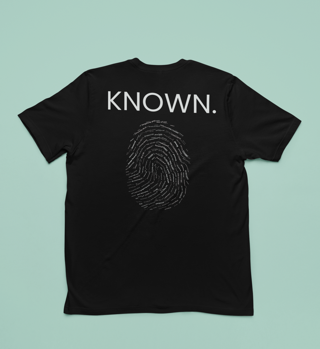 A black short-sleeve Christian t-shirt laid flat against a solid light mint green background. The back of the shirt features the word 'KNOWN.' with a fingerprint below inlaid with words of affirmation from God.