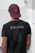 A young man viewed from behind wearing a maroon baseball cap that says Seed Sower Graphics and a black Christian Graphic t-shirt. The back of the t-shirt features the word 'KNOWN.' with a fingerprint inlaid with words of affirmation from the Bible