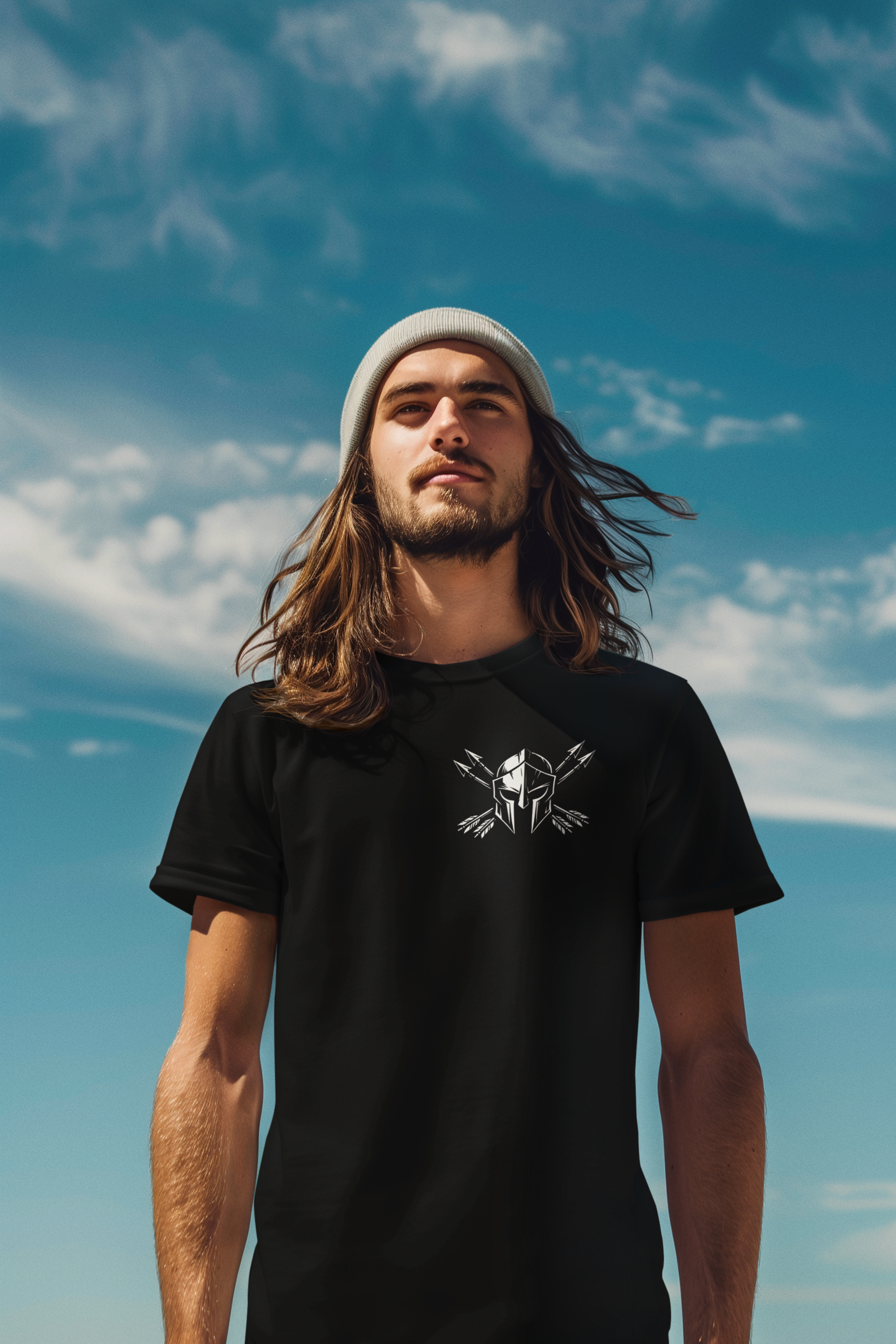 A young man wearing a black Like Arrow Christian Graphic t-shirt with a white graphic on the chest, a white beanie, and sunglasses, standing against a blue sky with clouds.