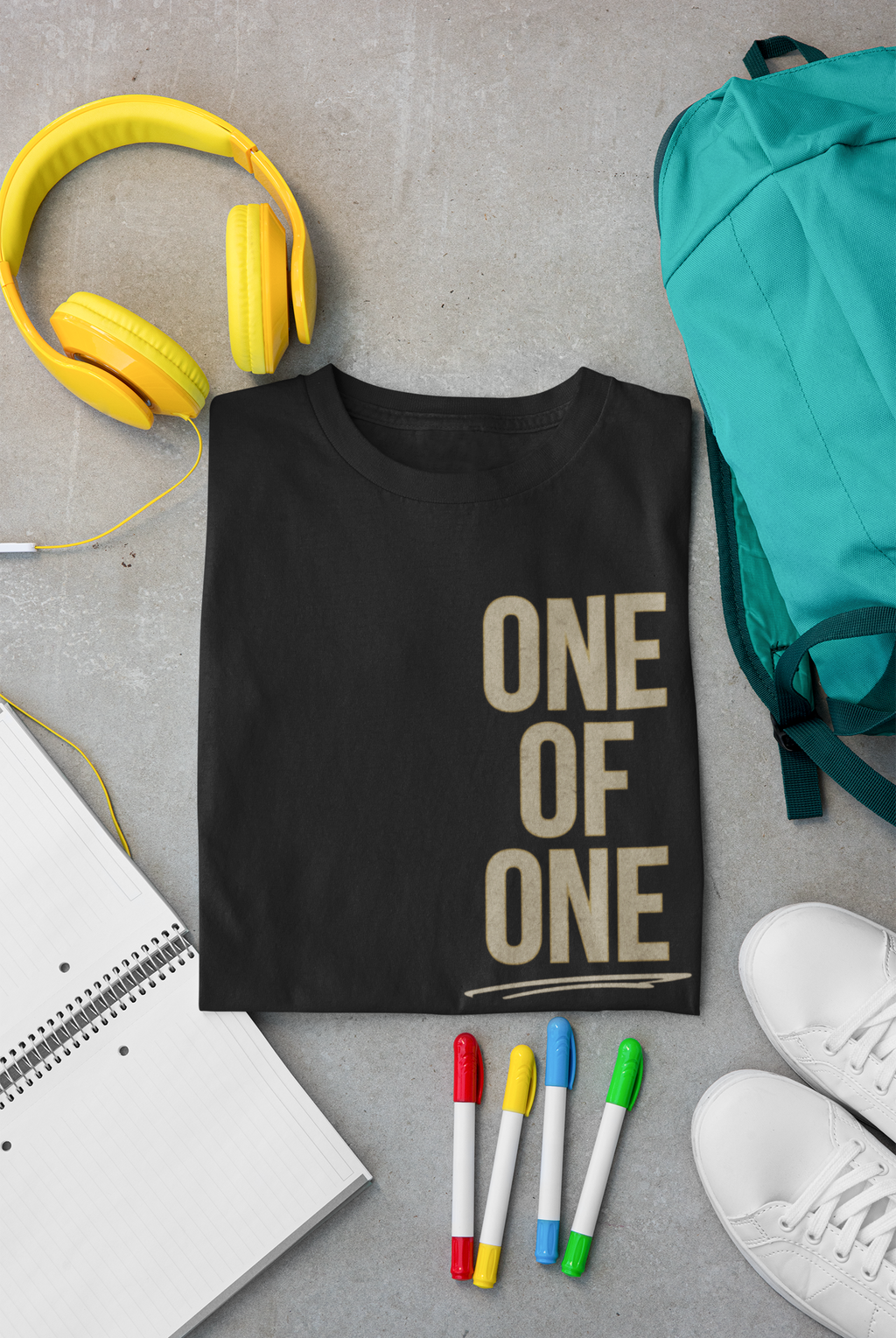 Image of a black folded Christian bible verse shirt that says 'One of One' vertically along the left side of the shirt in gold cream font. The shirt is surrounded by school related items such a yellow around the ear headphones, an aqua backpack, white sneakers, multicolored markers and an open notebook.