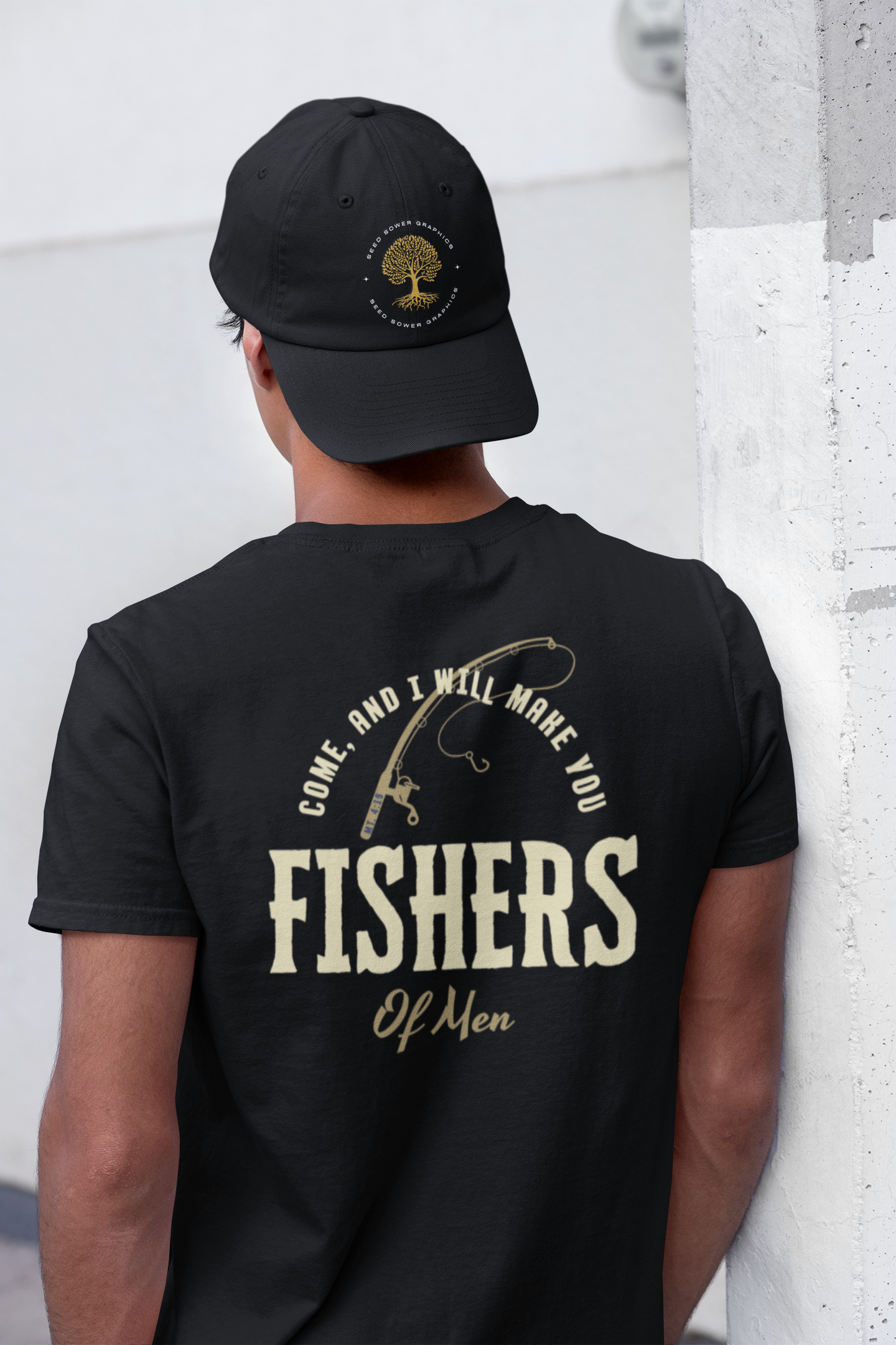 A man viewed from behind wearing a black Christian graphic t-shirt and a black baseball cap. The back of the t-shirt features a cream-colored graphic with a fishing rod, the text 'COME, AND I WILL MAKE YOU FISHERS Of Men', and a circular tree logo on the cap.