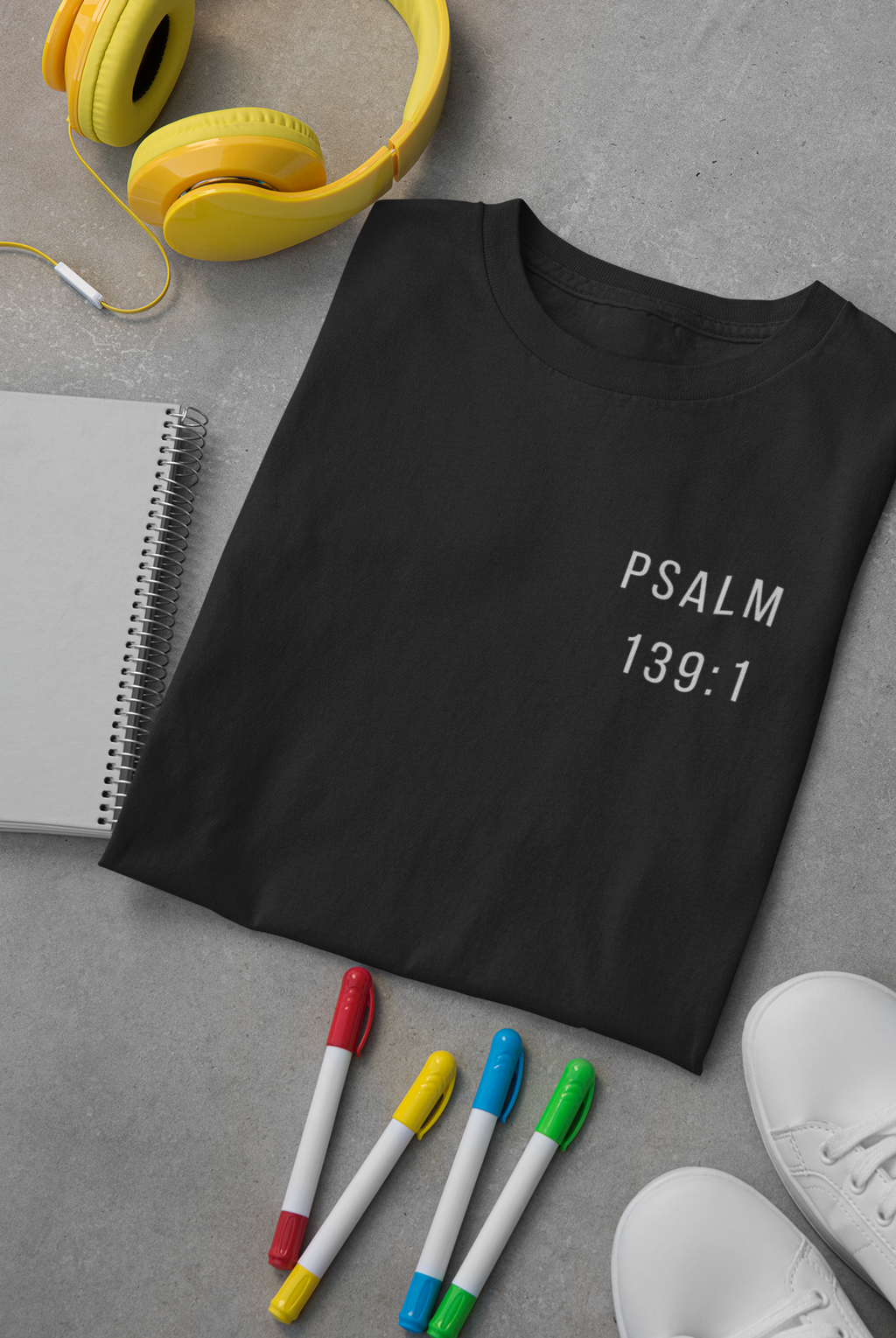 A black short-sleeve Christian T-shirt laid flat on a gray surface, printed with the text 'PSALM 139:1' in white on the left chest. Yellow headphones, a spiral notebook, four colored markers, and white sneakers are arranged around it as styled props.