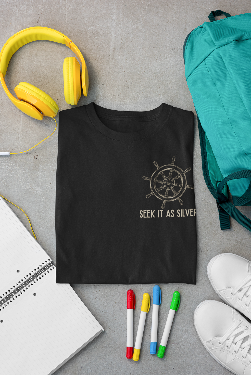 A folded black Christian t-shirt with a silver ship's wheel graphic and the text 'SEEK IT AS SILVER' is displayed in a flat-lay arrangement on a gray concrete background, surrounded by yellow headphones, a teal backpack, a notebook, colored markers, and white sneakers.
