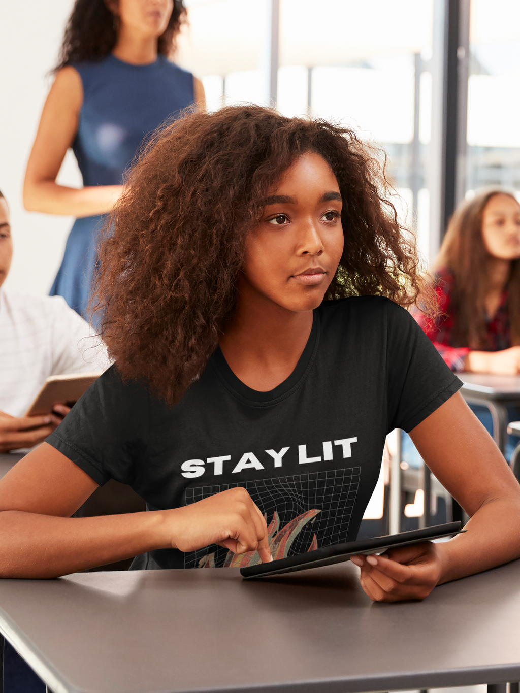 A girl with curly brown hair sits at a desk in a bright classroom or office, wearing a black Christian graphic T-shirt with the words 'STAY LIT' in white. She is holding a tablet, with other people blurred in the background.