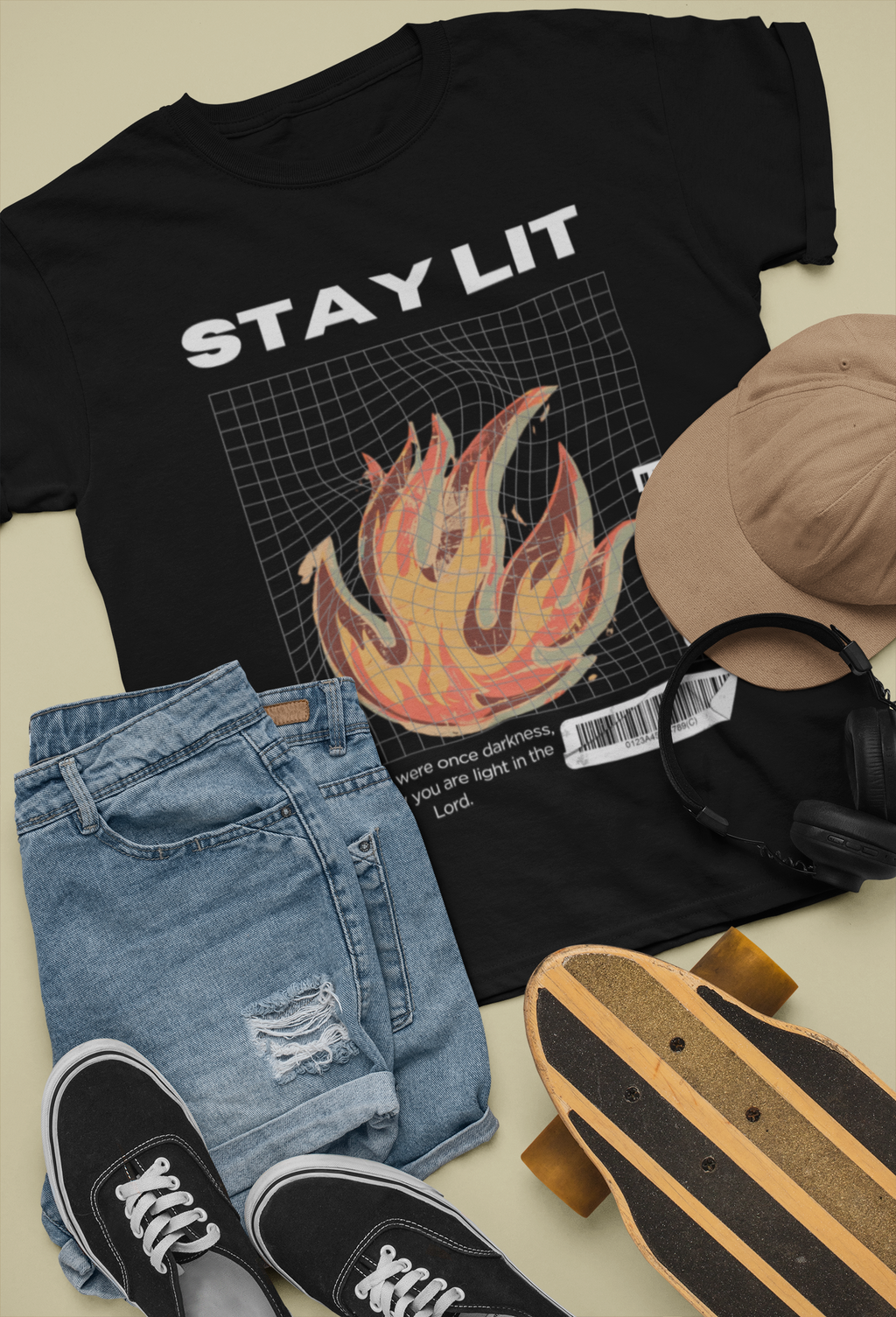 A styled flat-lay featuring a black Bible verse t-shirt with the text 'STAY LIT' and a flame design, paired with light-wash distressed jeans, black sneakers, a tan baseball cap, black headphones, and a skateboard on a light beige background