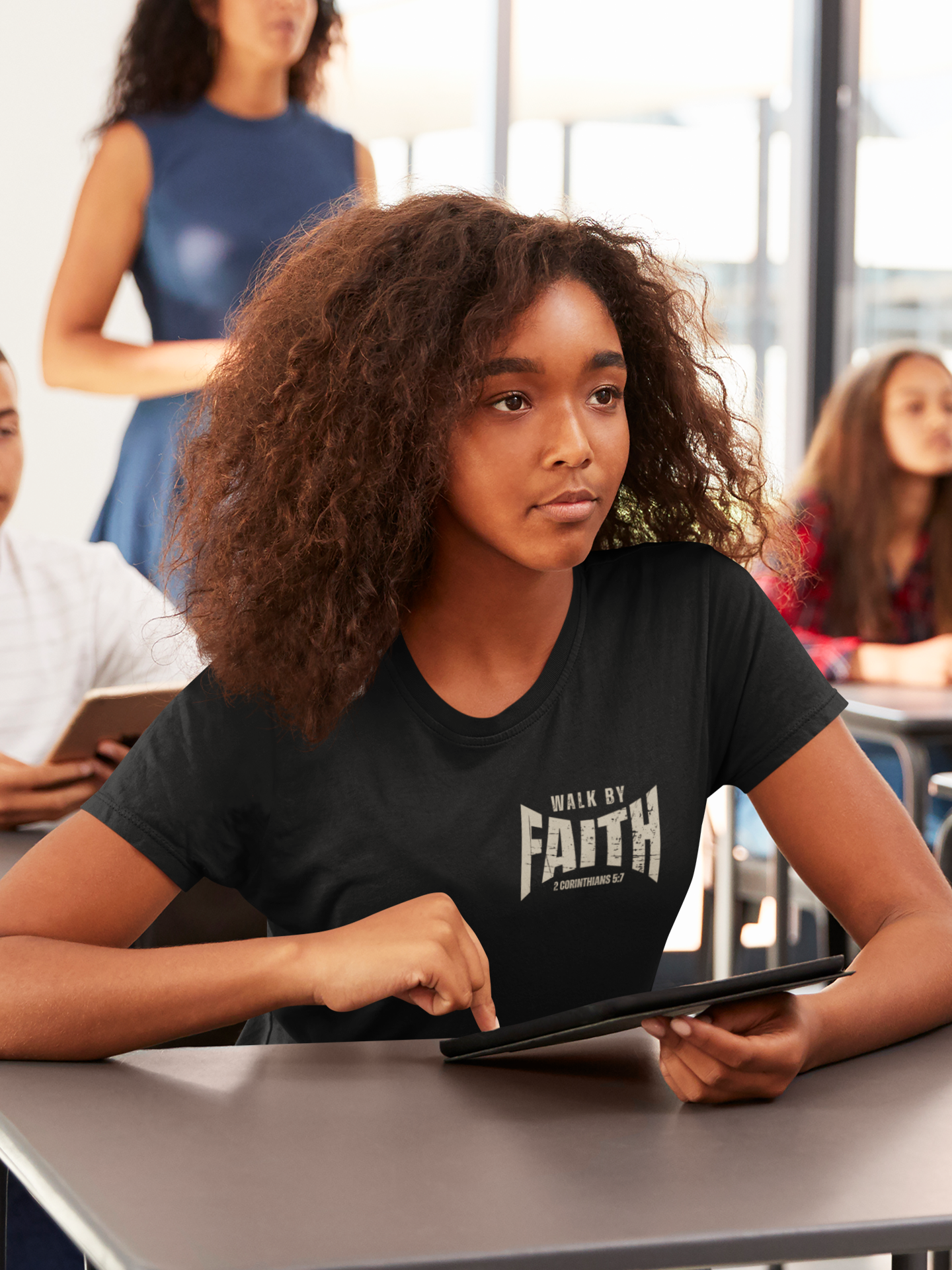 A woman with curly brown hair sits at a desk in a classroom setting, wearing a black Christian graphic T-shirt with the text 'Walk By Faith' in distressed cream lettering. She is holding a tablet, with other people blurred in the background.