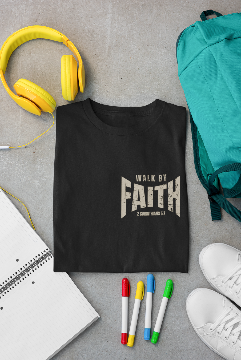 A folded black Christia T-shirt with a distressed white graphic reading 'WALK BY FAITH' and '2 Corinthians 5:7' is arranged in a styled flat lay on a gray surface, surrounded by yellow headphones, a teal backpack, a notebook, colored markers, and white sneakers.