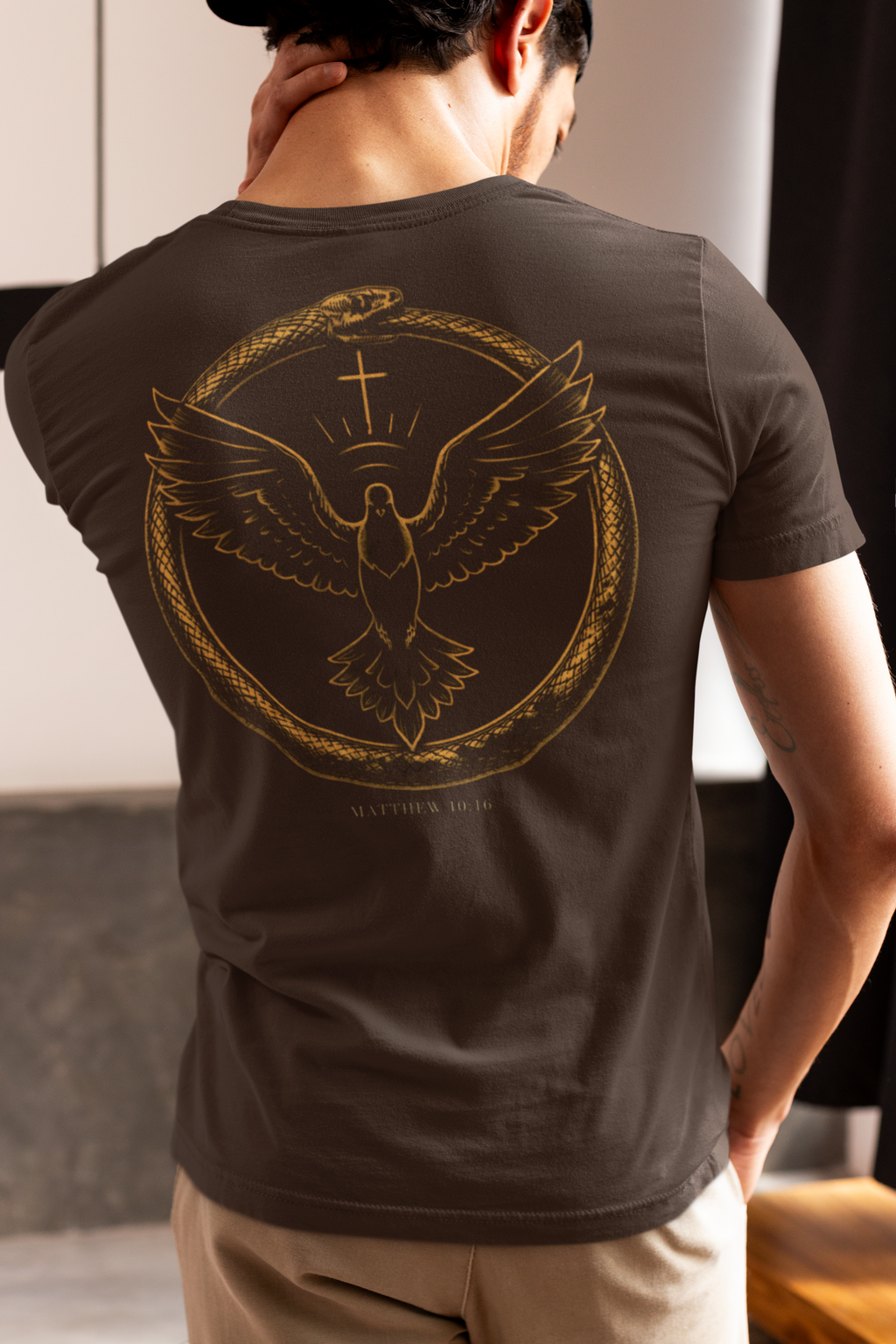 A man viewed from behind wearing a dark brown Christian graphic t-shirt with a large gold graphic of a dove with wings spread inside a circular frame made of a serpent with the verse 'Matthew 10:16' below it, on a neutral indoor background.