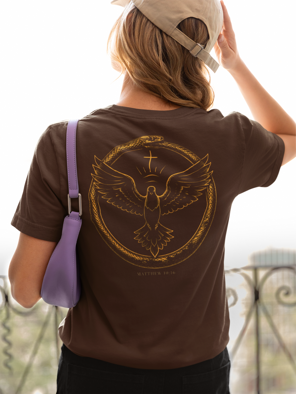 Person wearing a brown Christian t-shirt with a gold dove design with the word 'Matthew 10;16' below it, holding a beige cap and purple bag.
