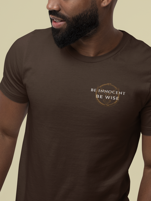 A close-up of a man wearing a dark brown Christian graphic t-shirt with a gold and white graphic on the left chest that reads 'BE INNOCENT BE WISE' inside a circular emblem, set against a solid beige background.