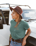 A woman wearing a dusty green Christian graphic t-shirt with a brown 'SS seed sower' graphic, paired with a brown felt hat and blue jeans, posing next to a white Jeep outdoors.