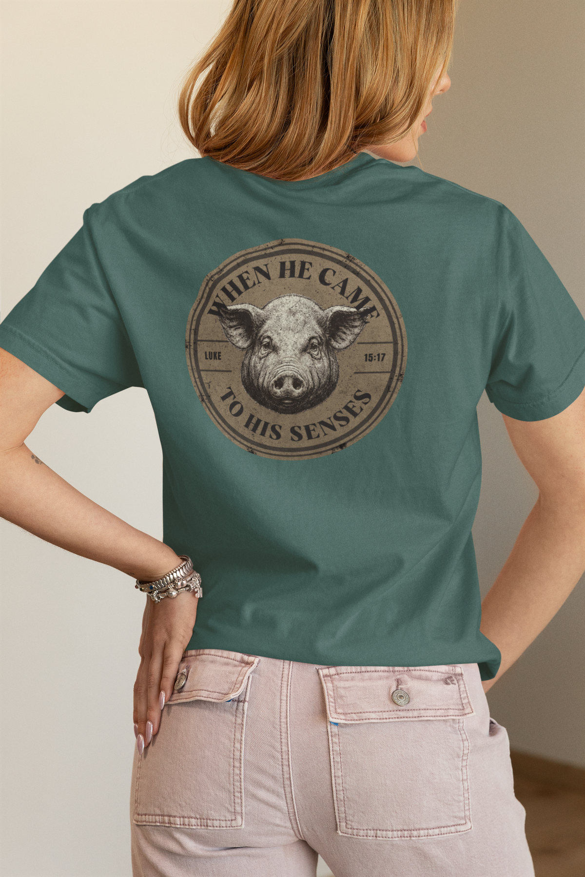 A woman shown from the back wearing a teal green Chrisitan bible verse t-shirt with a circular graphic of a pig's face and the text 'wHEN HE CAME TO HIS SENSES' on the back, paired with light pink jeans.