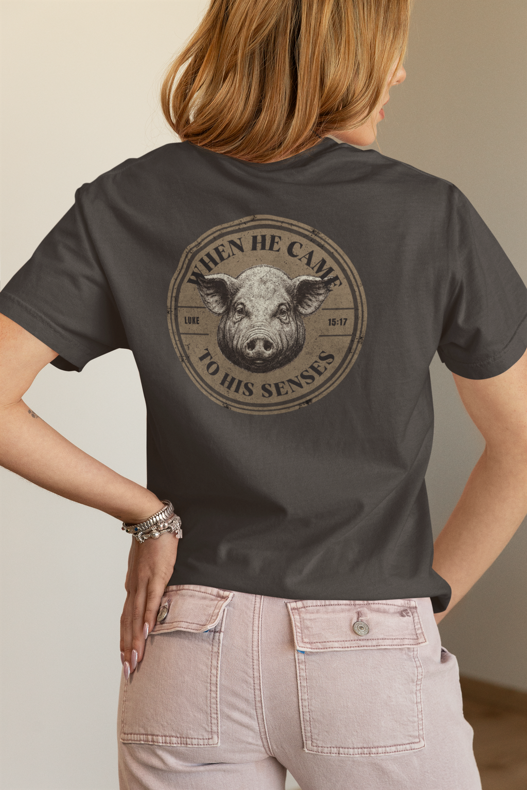 A woman shown from the back wearing a dark charcoal gray Christian t-shirt with a circular graphic of a pig's face and the text 'THEN HE CAME TO HIS SENSES' on the back, paired with light pink jeans.
