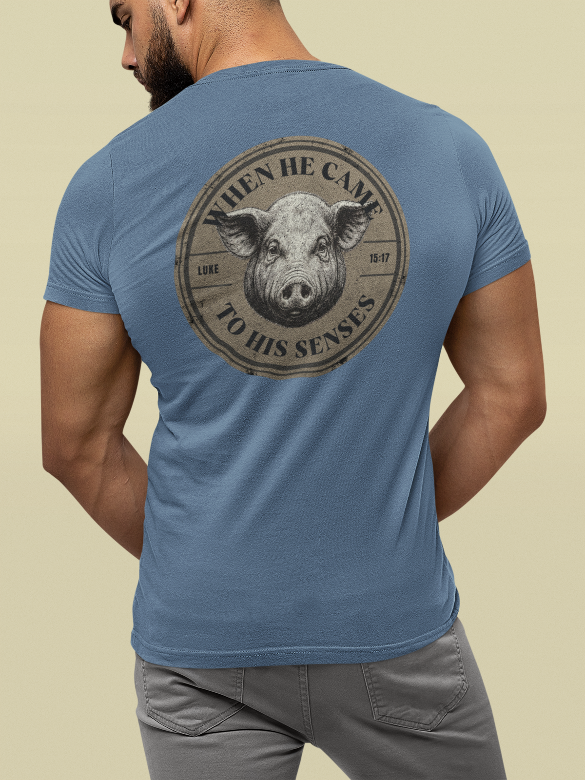 A man wearing a blue crewneck Christian t-shirt, viewed from the back. The shirt features a large circular graphic print on the back with a pig's face and the text 'WHEN HE CAME TO HIS SENSES'.