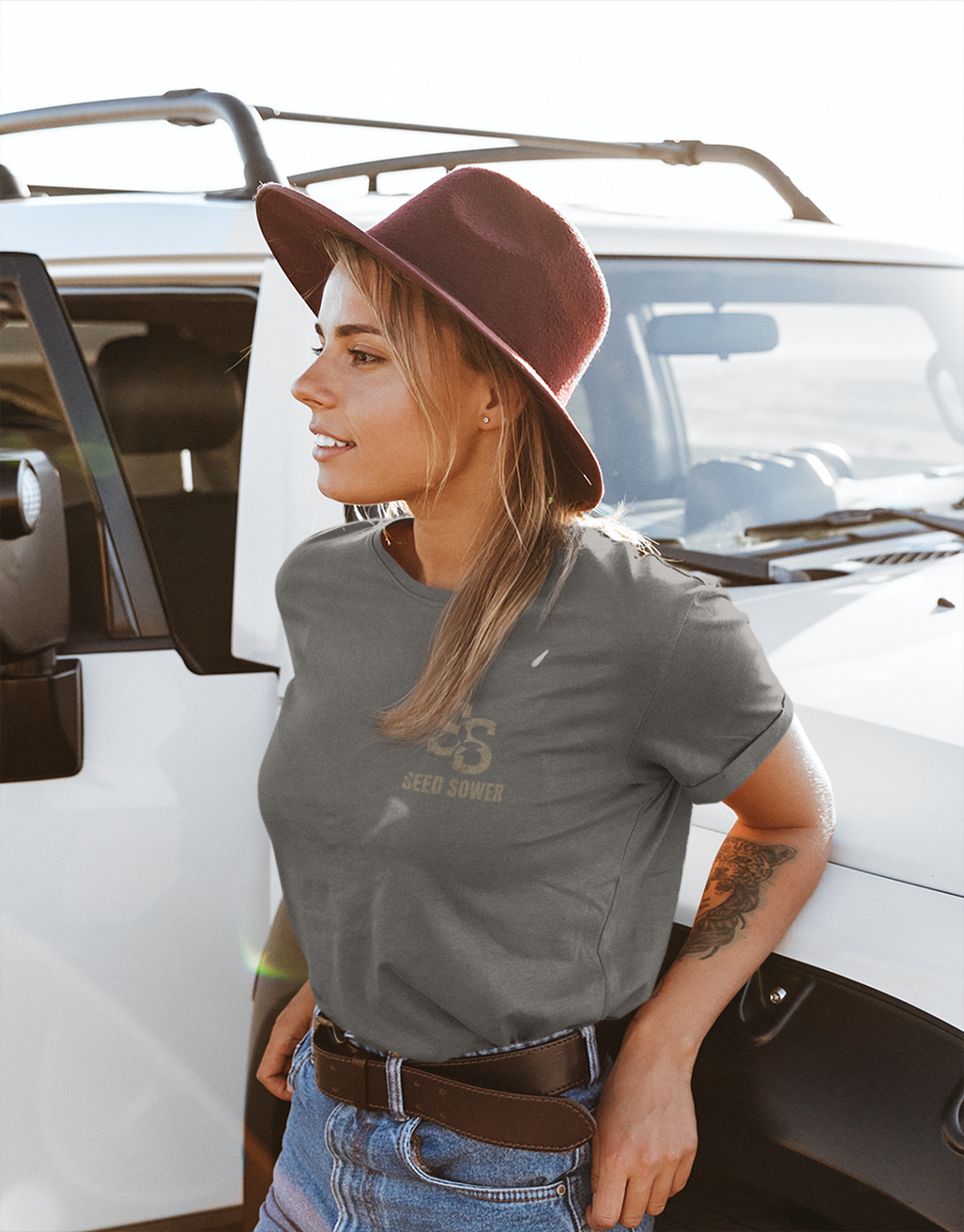 A woman wearing a green Christian graphic t-shirt with a brown SS seed sower print and a brown felt hat, posing next to a white vehicle.