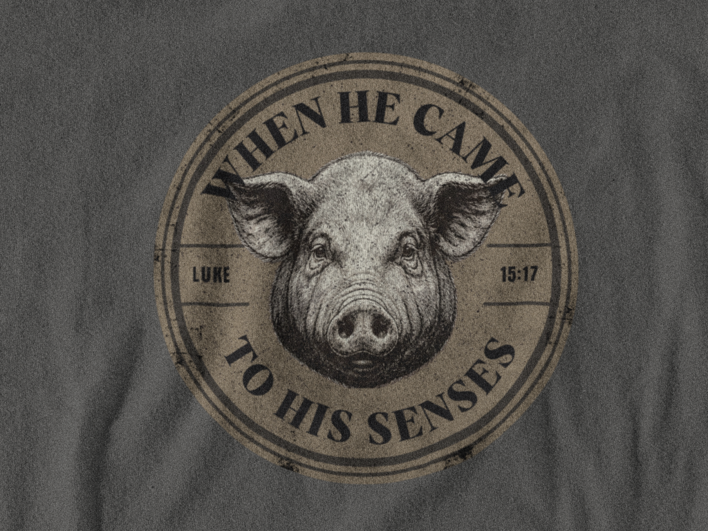 A close-up of a dark gray christian graphic shirt featuring a circular distressed graphic print with a pig's face in the center and the text 'WHEN HE CAME TO HIS SENSES' and 'LUKE 15:17'.