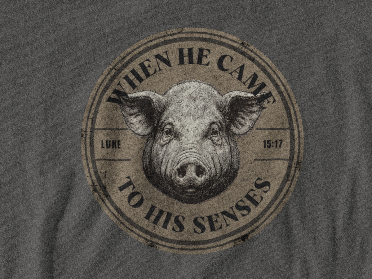 A close-up of a dark gray christian graphic shirt featuring a circular distressed graphic print with a pig's face in the center and the text 'WHEN HE CAME TO HIS SENSES' and 'LUKE 15:17'.