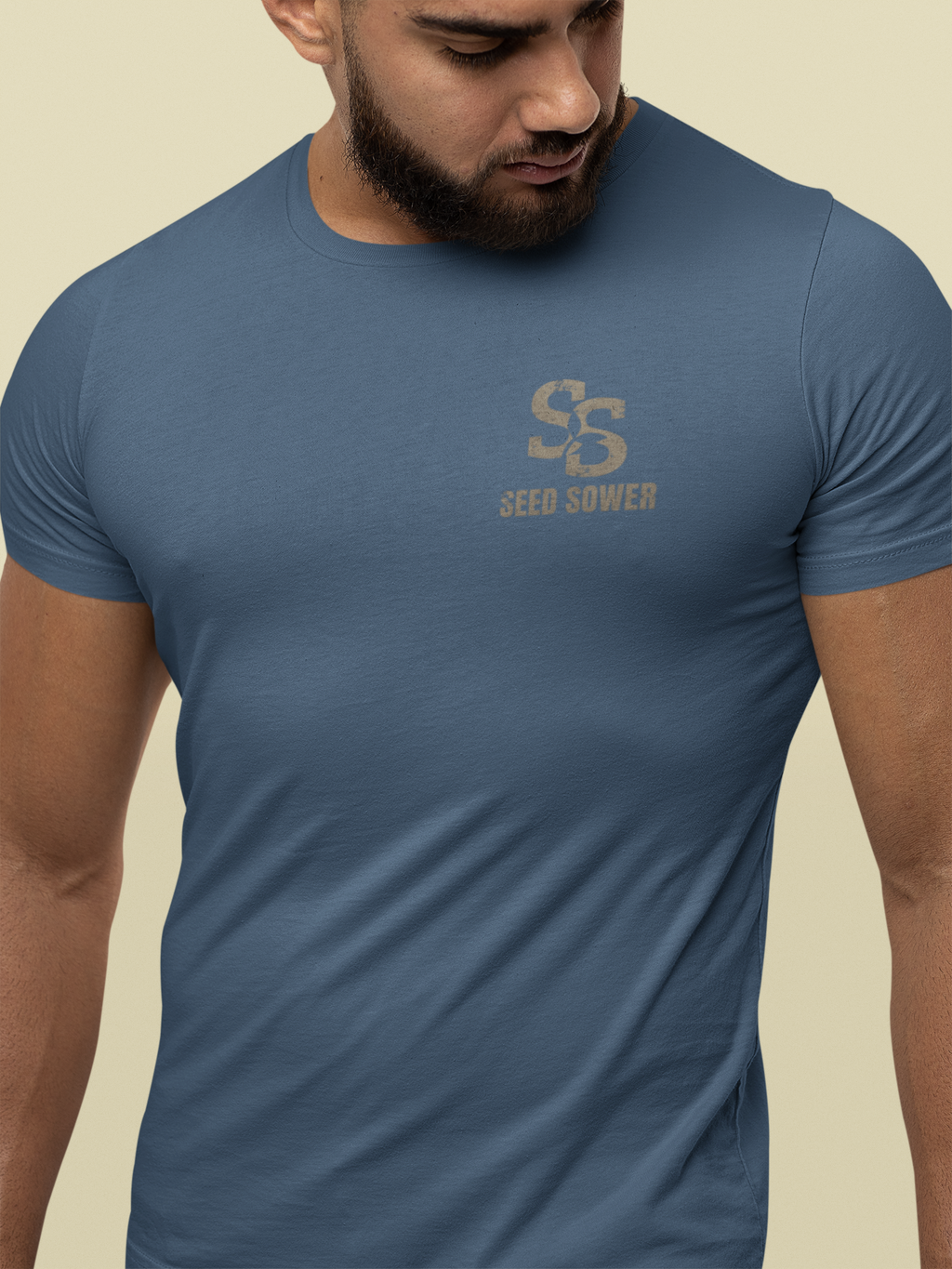 A man wearing a slate blue crewneck Christian graphic t-shirt with a distressed tan 'Seed Sower' logo on the chest, set against a solid beige background.