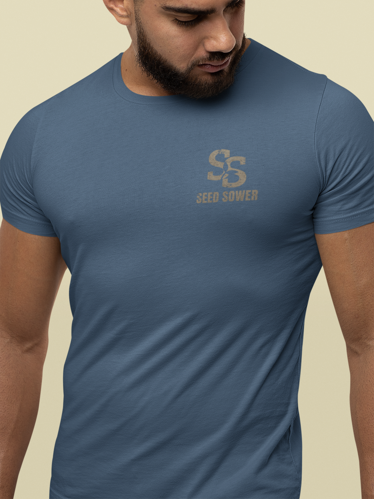 A man wearing a slate blue crewneck Christian graphic t-shirt with a distressed tan 'Seed Sower' logo on the chest, set against a solid beige background.
