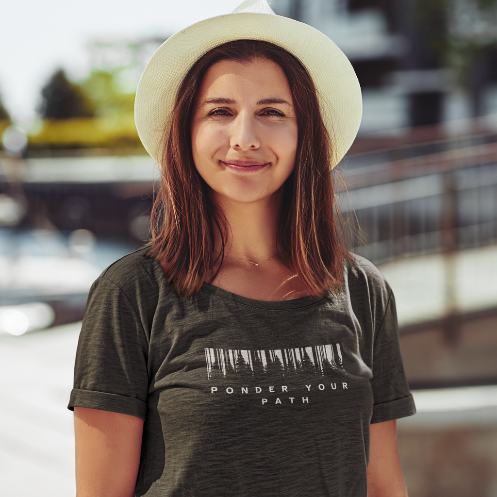 A woman wearing a dark heather Christian graphic t-shirt with a barcode graphic and the text 'PONDER YOUR PATH' underneath, paired with a cream-colored wide-brim hat, set against a blurred outdoor background.