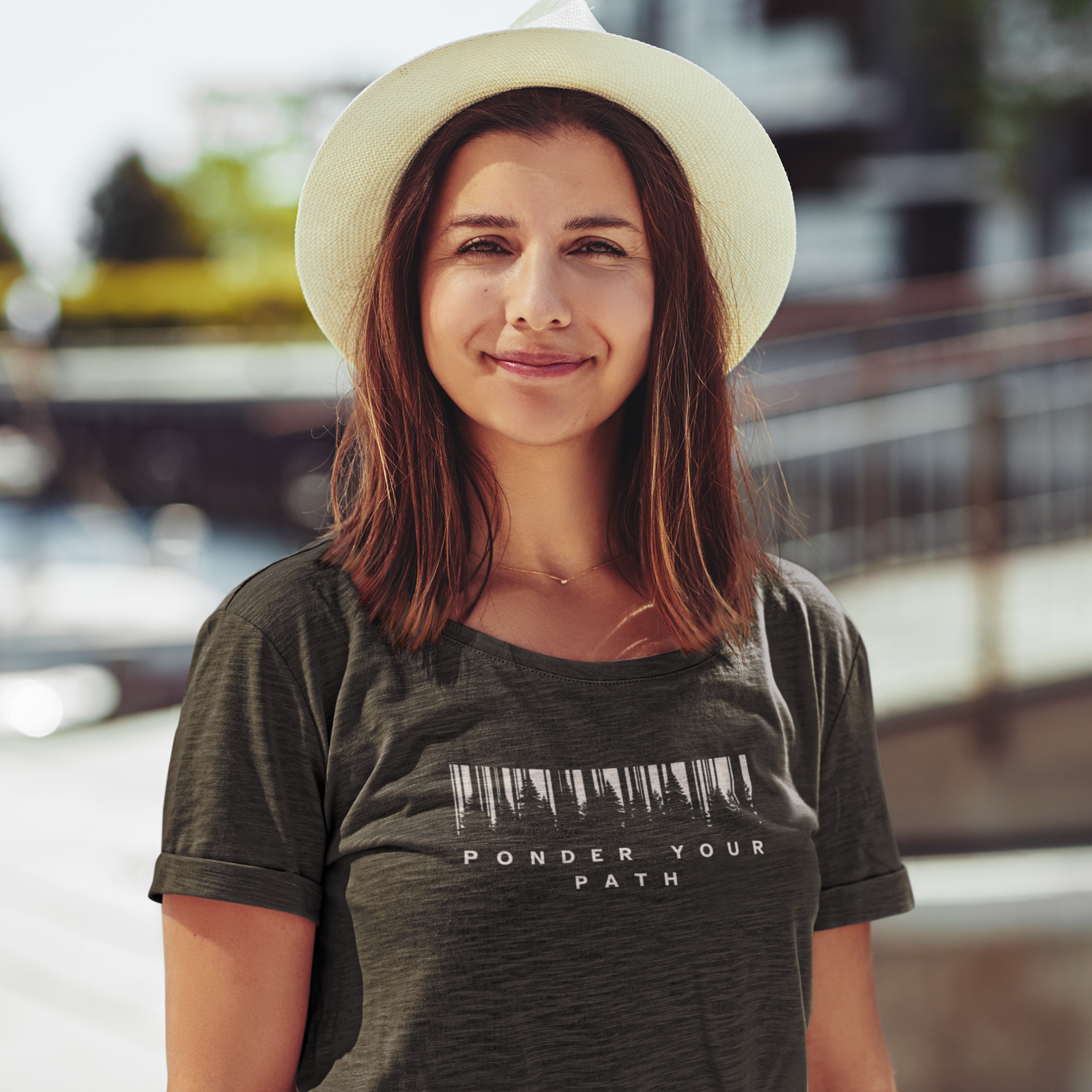A woman wearing a dark heather Christian graphic t-shirt with a barcode graphic and the text 'PONDER YOUR PATH' underneath, paired with a cream-colored wide-brim hat, set against a blurred outdoor background.