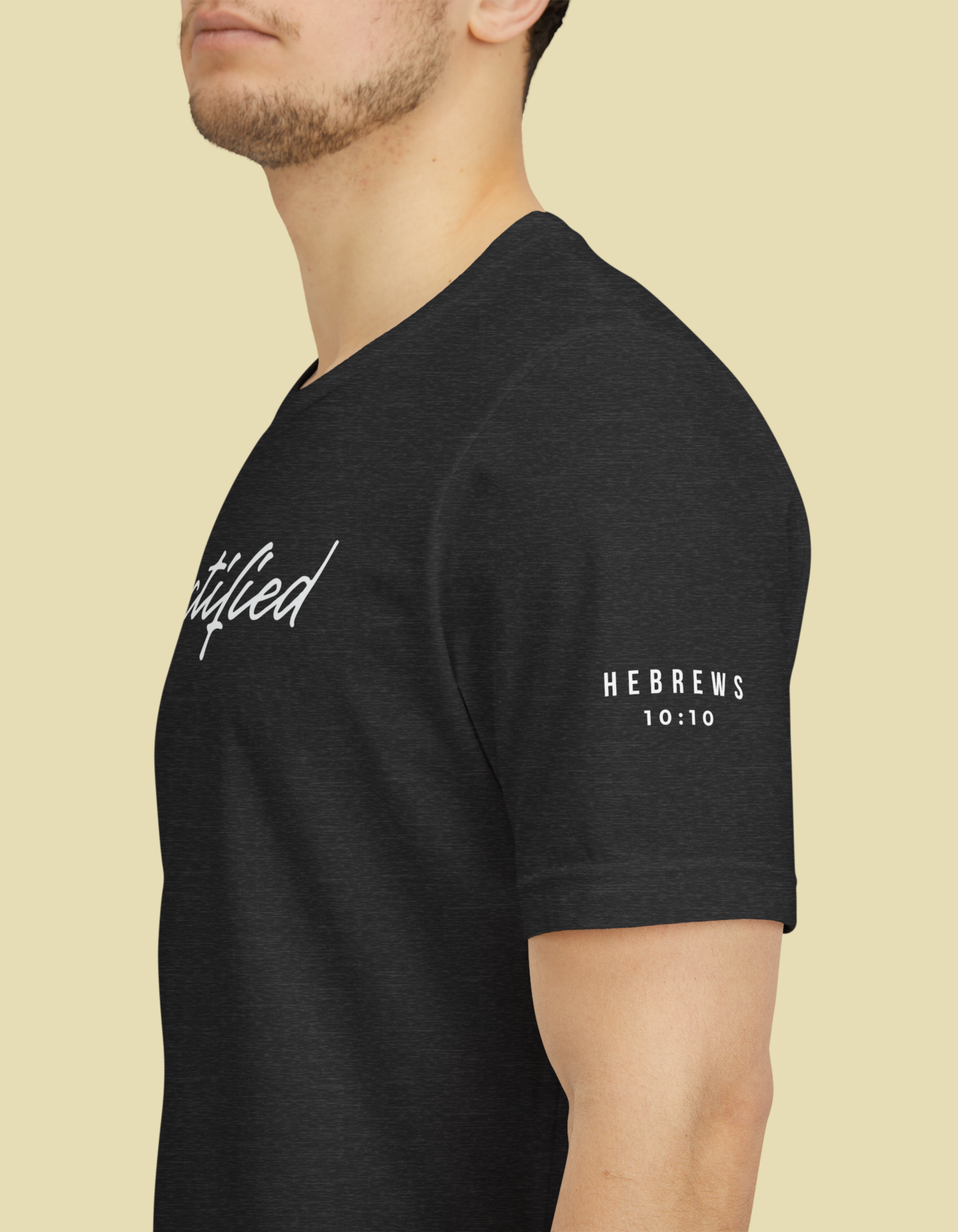 A man wearing a dark heather Christian bible verse t-shirt with white printed text 'Sanctified'on the chest and 'Hebrews 10:10' on the left sleeve, shown in a side profile against a solid light beige background.
