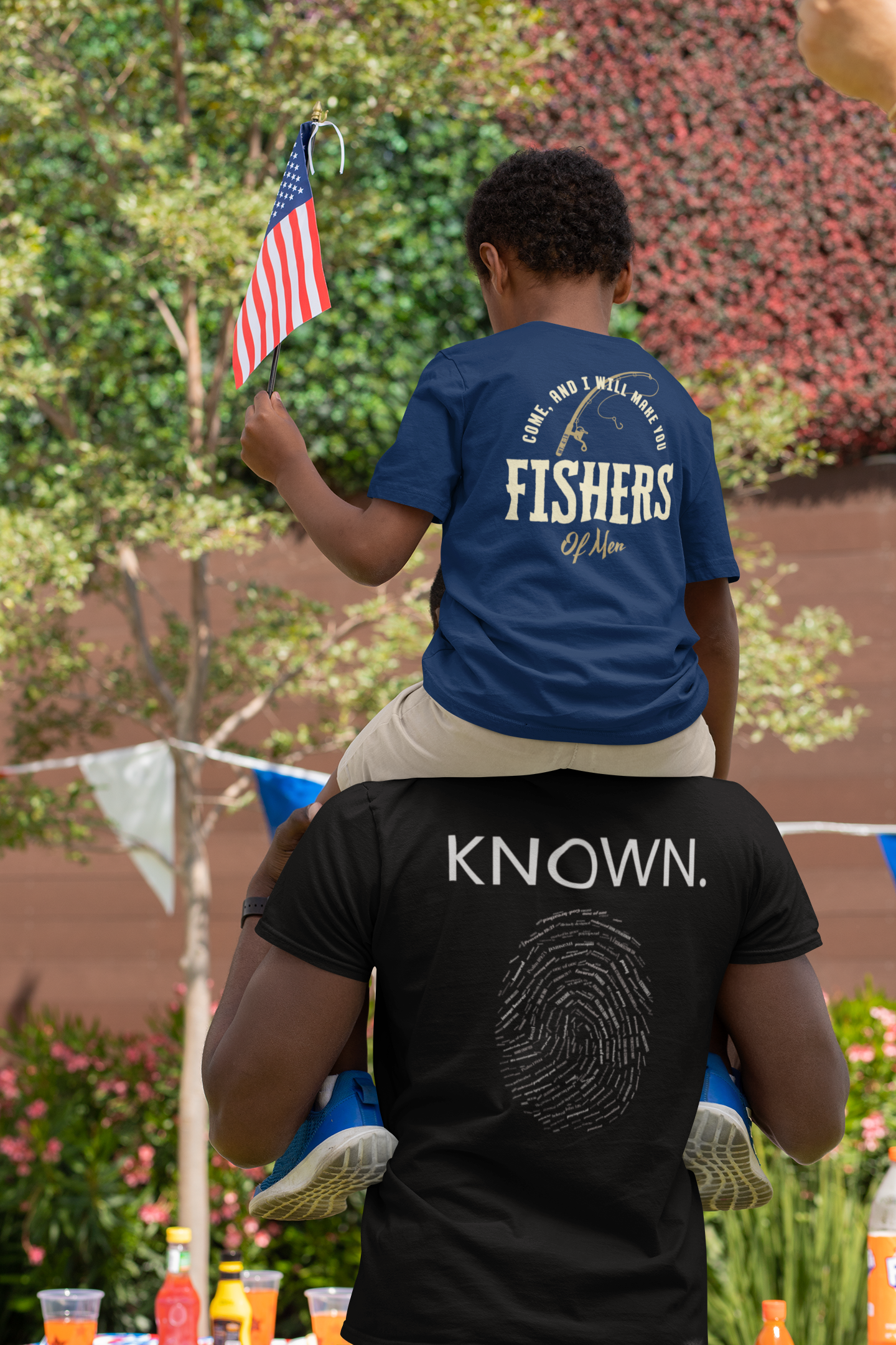 A father and son wearing meaningful Christian t-shirts with 'KNOWN.' and a fisherman logo on the back, photographed from behind in an outdoor setting.