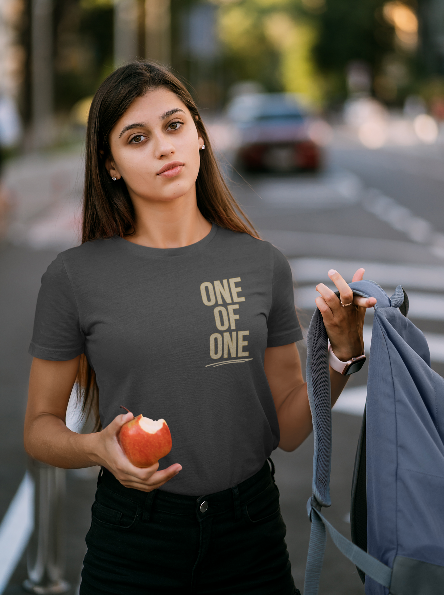 A young female student looking into the camera putting on a blue backpack with a bitten apple in the other hand. She is wearing a gray Christian graphic tee with cream colored words affixed vertically along the left side of her shirt with the words, 'One of One'. 