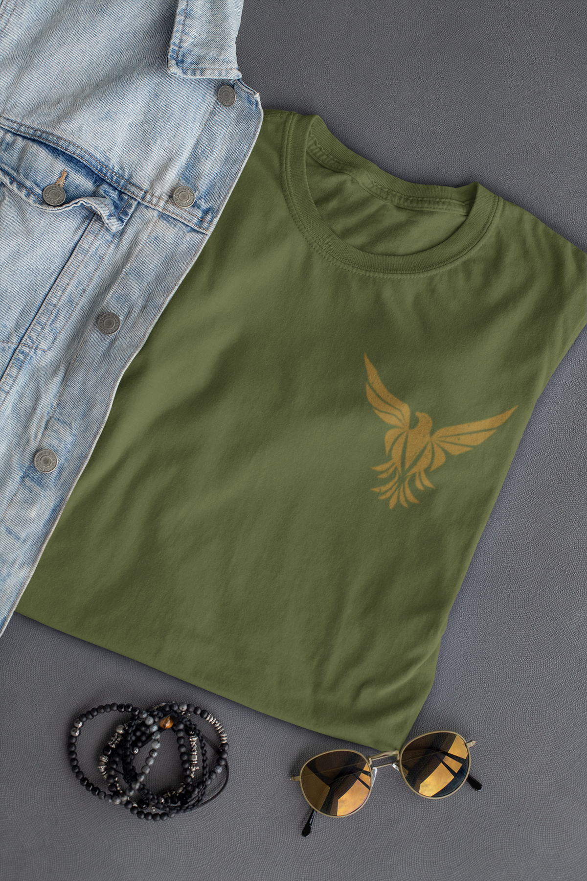 A military green crewneck faith t-shirt with a gold eagle graphic on the left chest, styled in a flat-lay arrangement with a light blue denim shirt, black beaded bracelets, and sunglasses on a dark gray background.
