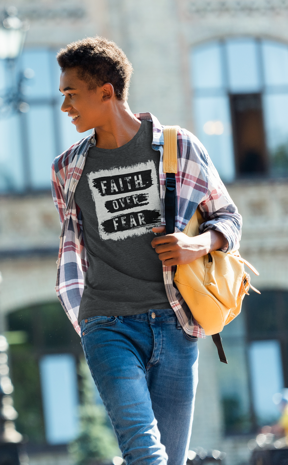 A young man walking outdoors wearing a dark gray Christian graphic t-shirt with 'FAITH OVER FEAR' printed in white, layered under an open red and blue plaid flannel shirt, paired with blue jeans and a yellow backpack.