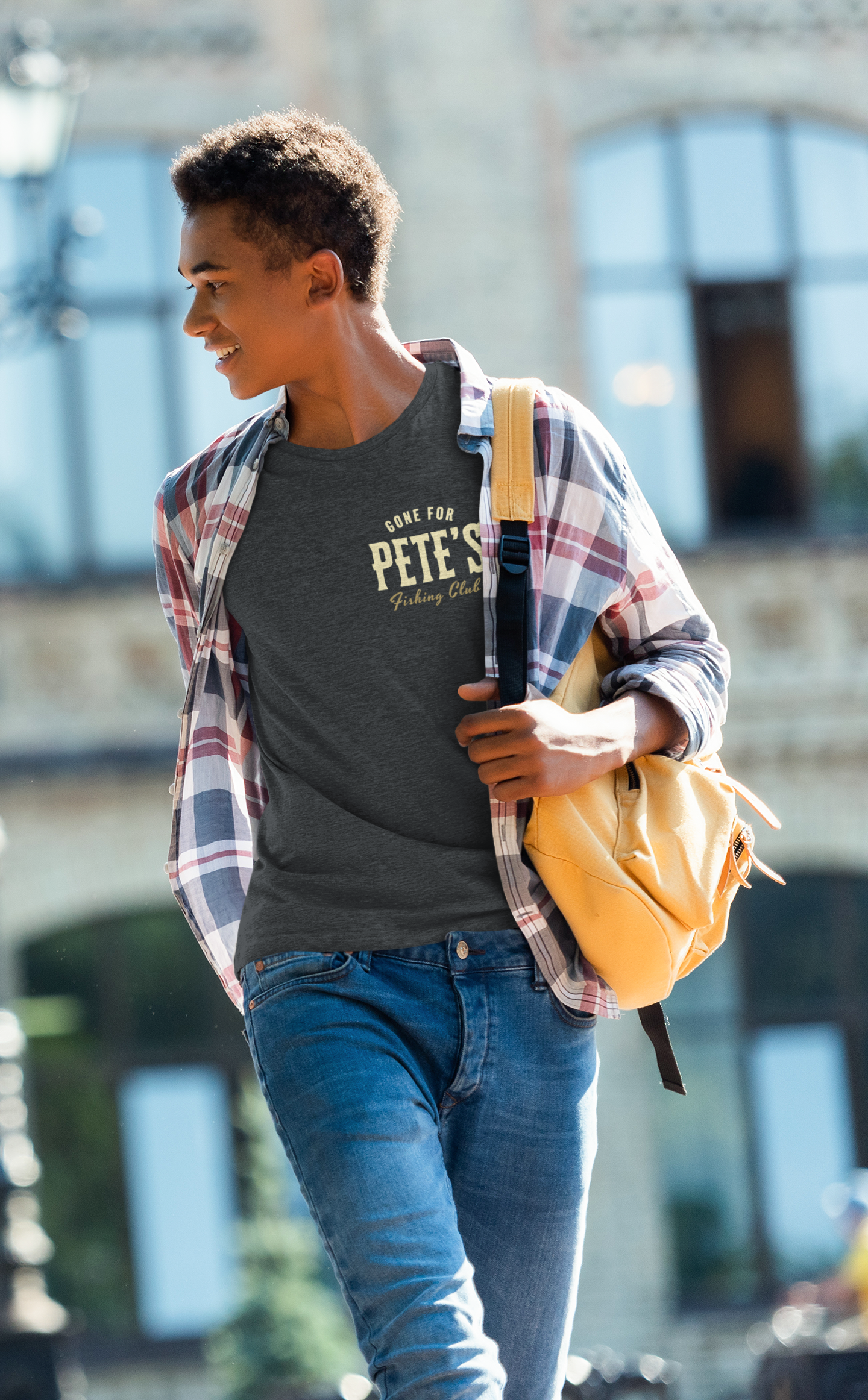 A young man walking outdoors wearing a dark gray Christian graphic t-shirt that says 'GONE FOR PETE'S FISHING CLUB' under an open red and blue plaid flannel shirt, blue jeans, and carrying a yellow backpack.