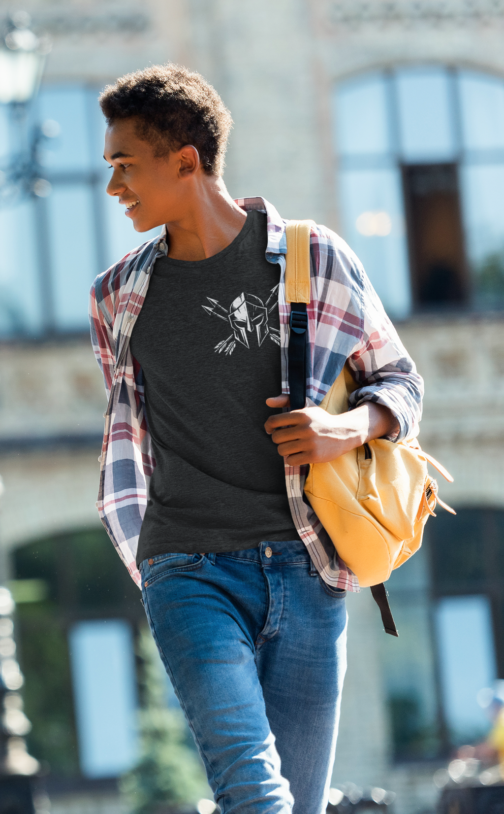 A young man walking outdoors wearing a dark gray Christian graphic t-shirt under an open red and blue plaid flannel shirt, blue jeans, and carrying a yellow backpack.