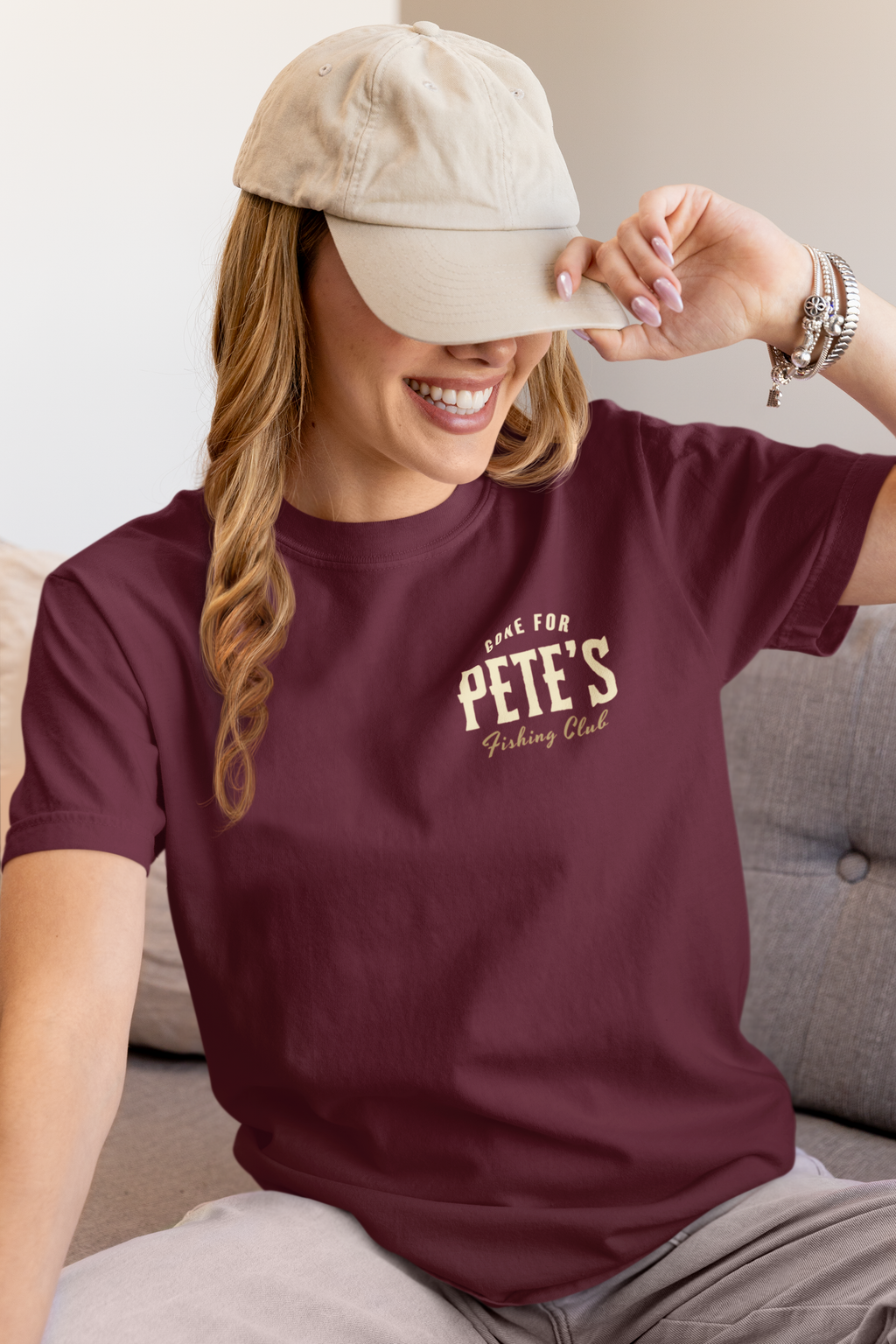 A woman wearing a maroon Christian graphic t-shirt with a cream graphic that reads 'Gone FOR PETE'S Fishing Club' and a beige baseball cap, sitting on a gray couch.
